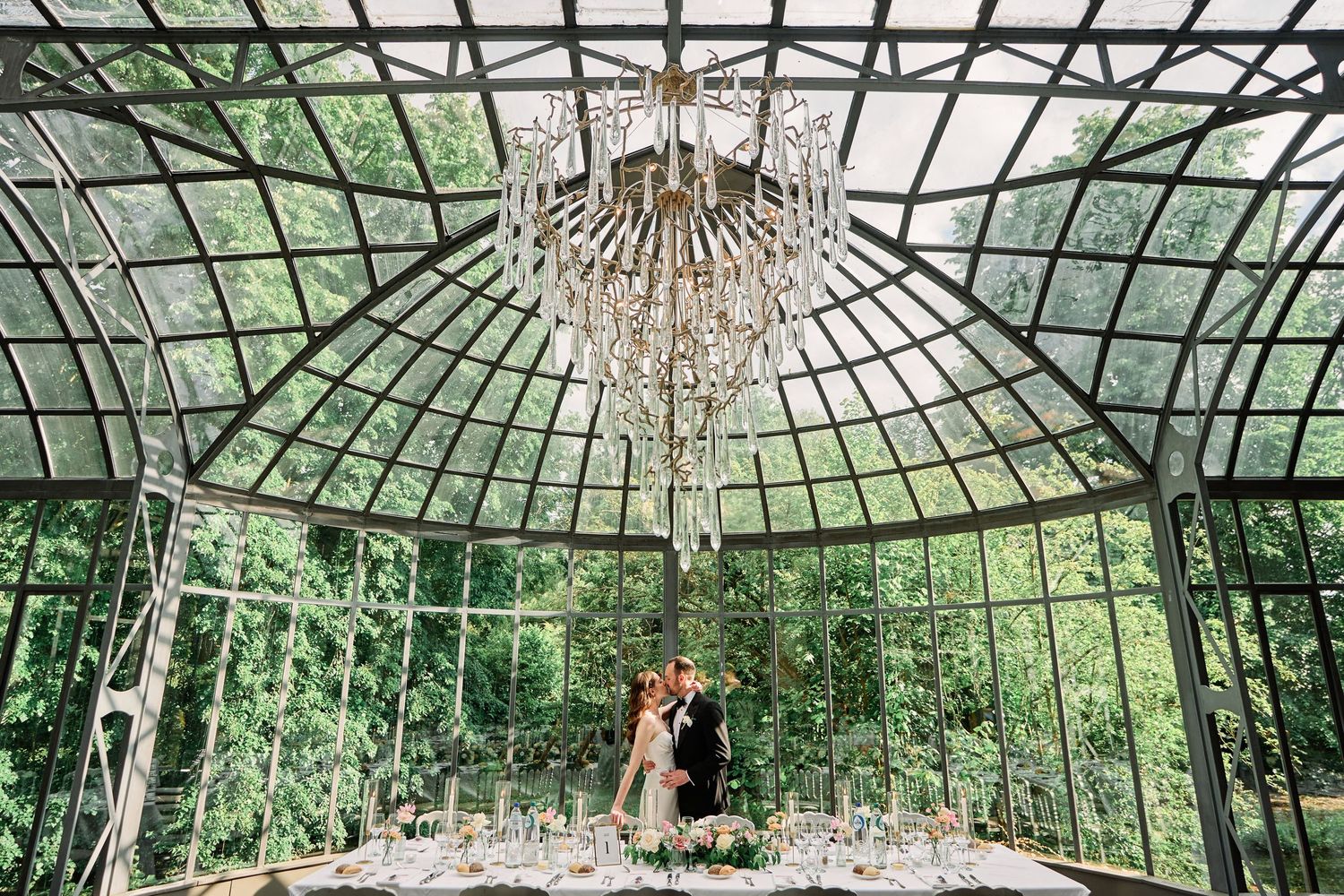 Crystal chandeliers hang from a glass conservatory ceiling with lush greenery and natural light streaming in.