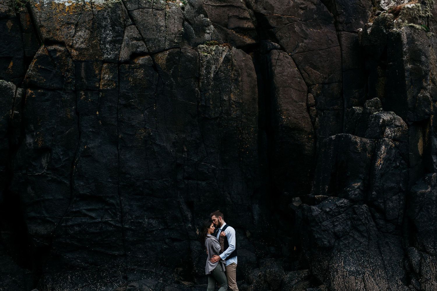 A person stands against a dark rocky cliff face wearing a backpack and climbing gear.