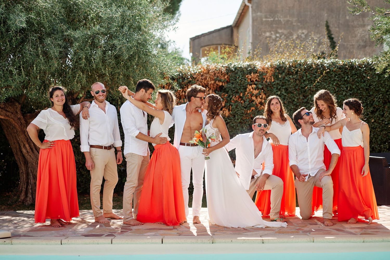 Wedding party in coral dresses and white suits pose together in an outdoor garden setting.