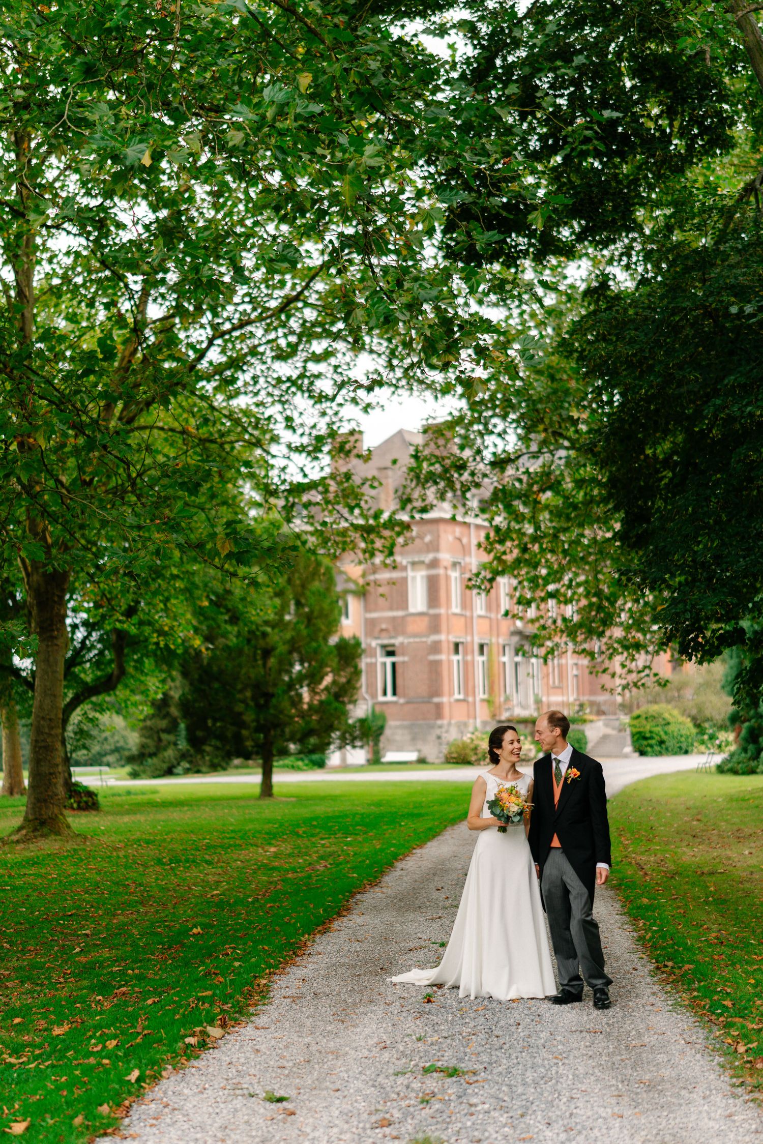 A couple walks along a tree-lined gravel path on the grounds of a historic brick mansion on their wedding day.