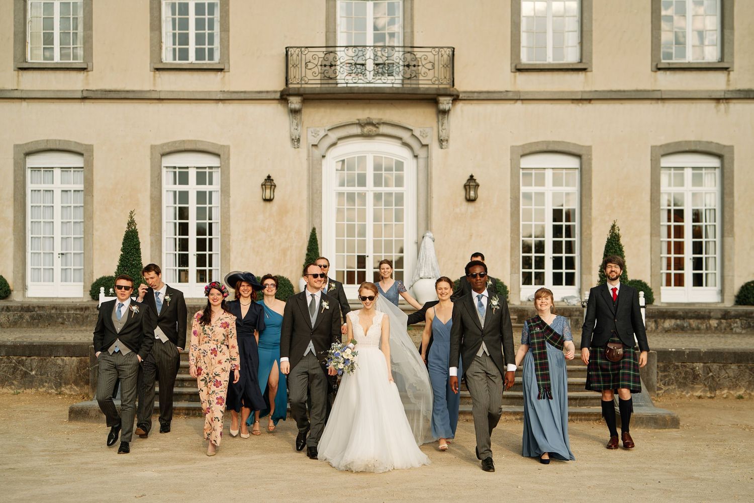 A wedding party celebrates outside an elegant French-style mansion with tall windows and classic architecture.