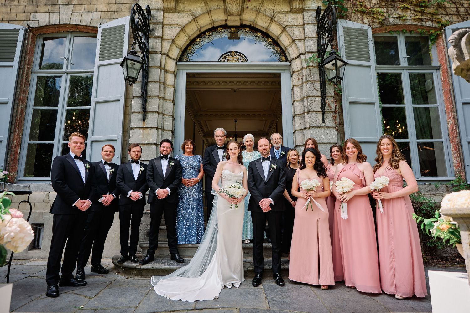 Wedding party poses together in front of a stone building with elegant arched windows and doorways.