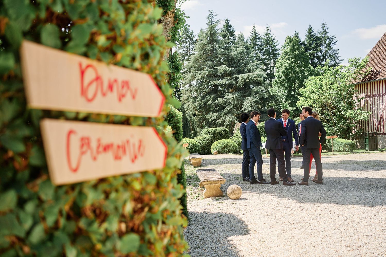 Wooden directional signs with orange text mounted on posts along a garden pathway with evergreen trees in background.