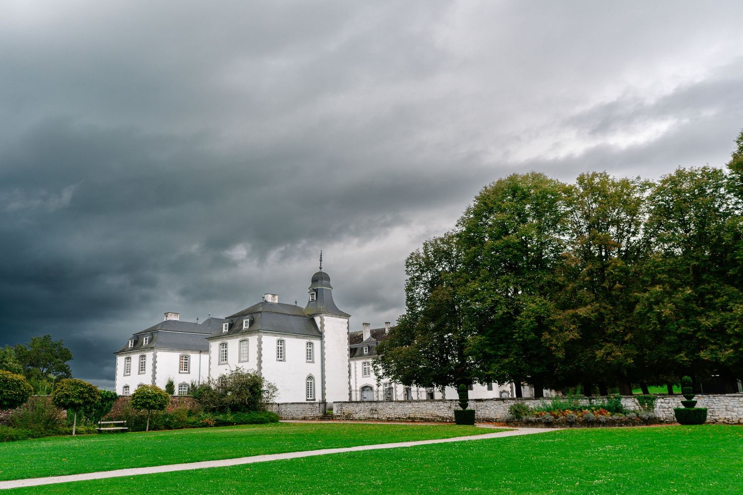 White church building with spire stands against dark stormy clouds and green lawn in a dramatic countryside setting.