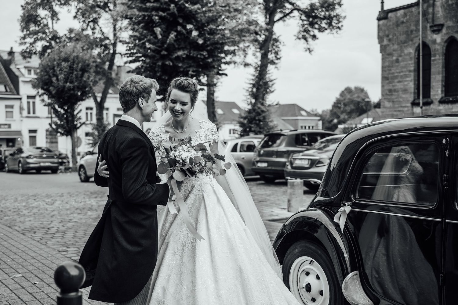 A vintage black and white wedding scene with a classic car parked outside a church on a tree-lined street.