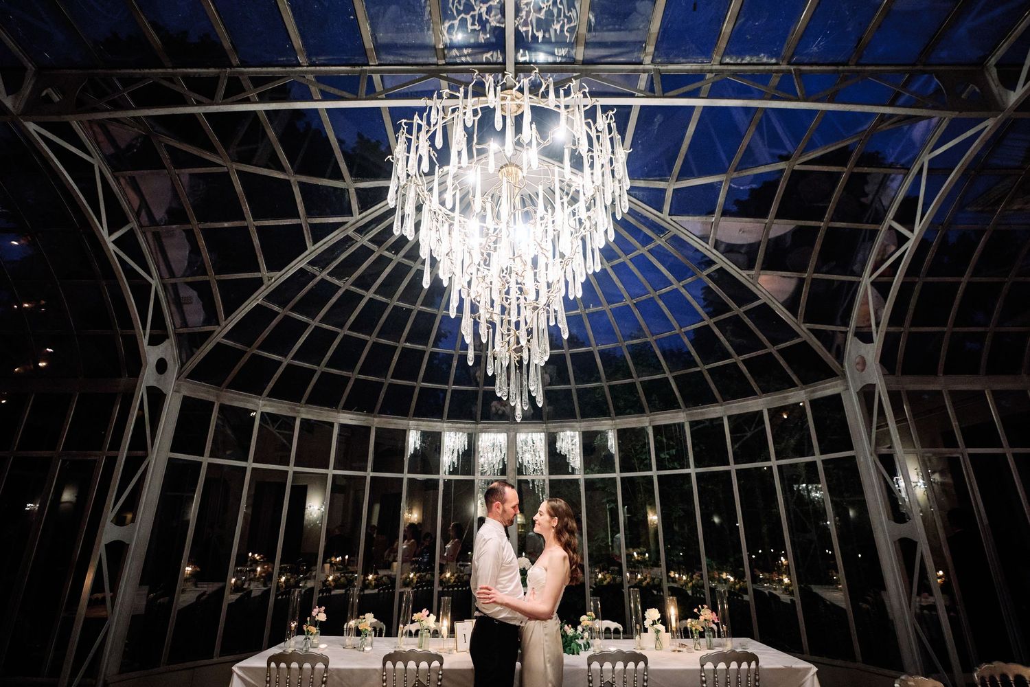 A grand crystal chandelier hangs from an ornate glass ceiling in an elegant ballroom setting.