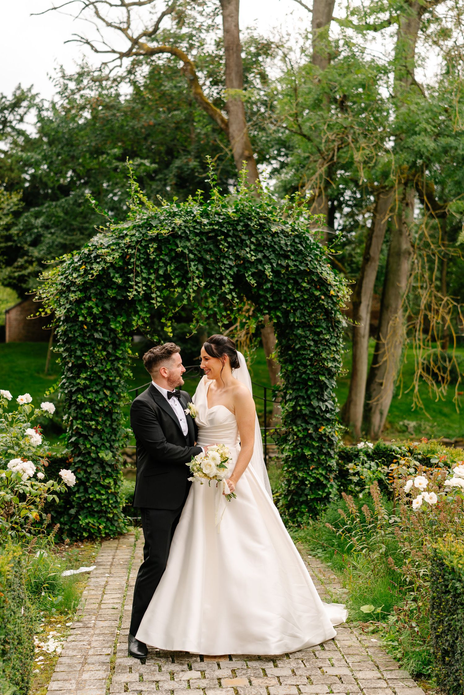 A newlywed couple shares romantic moments in a lush garden setting with a stone pathway and blooming flowers.