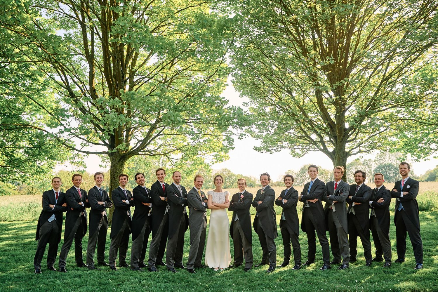 Wedding party poses under lush green trees for outdoor group photos during summer celebration.