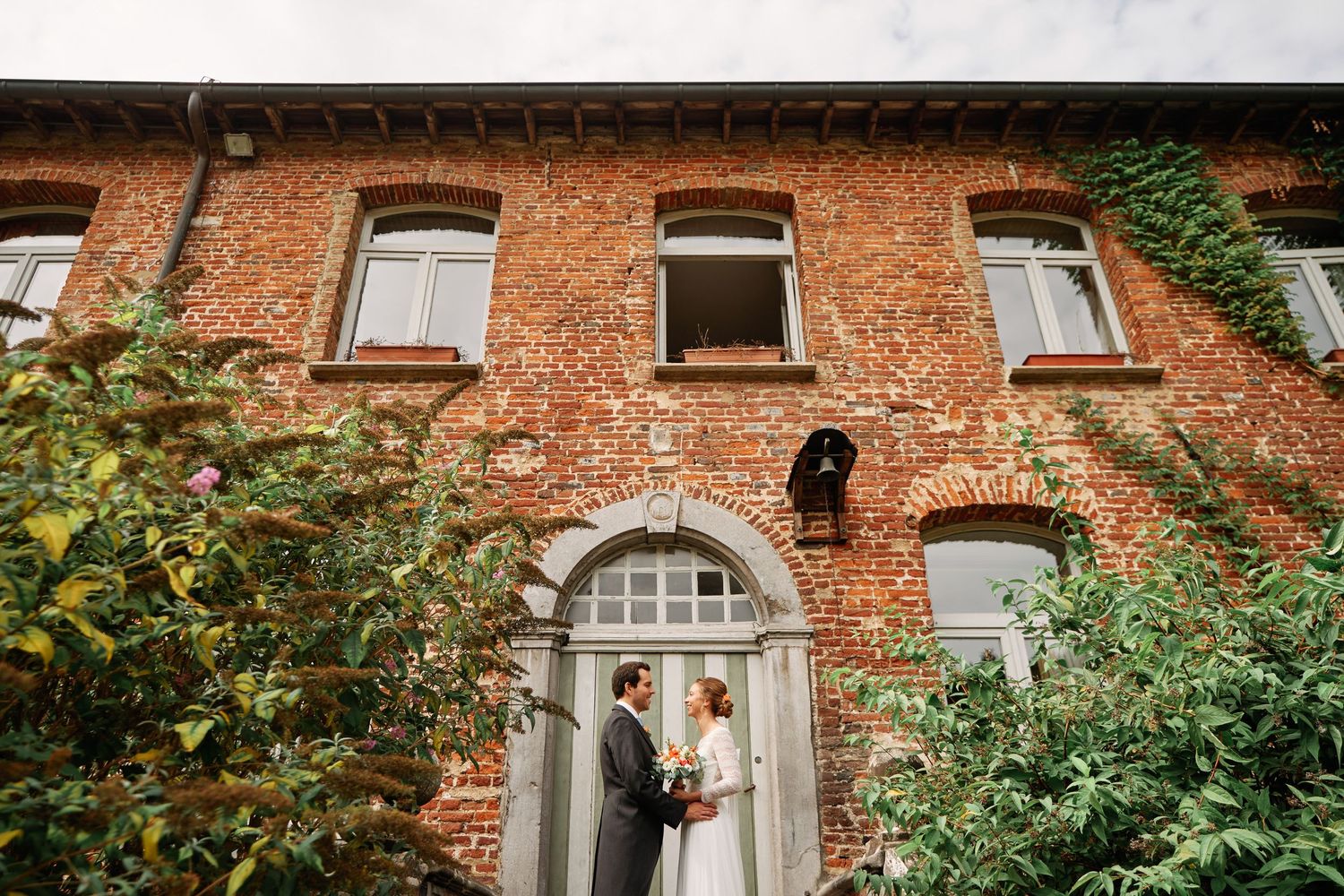 A couple stands in front of a rustic brick building entrance with arched doorway and greenery growing on the facade.