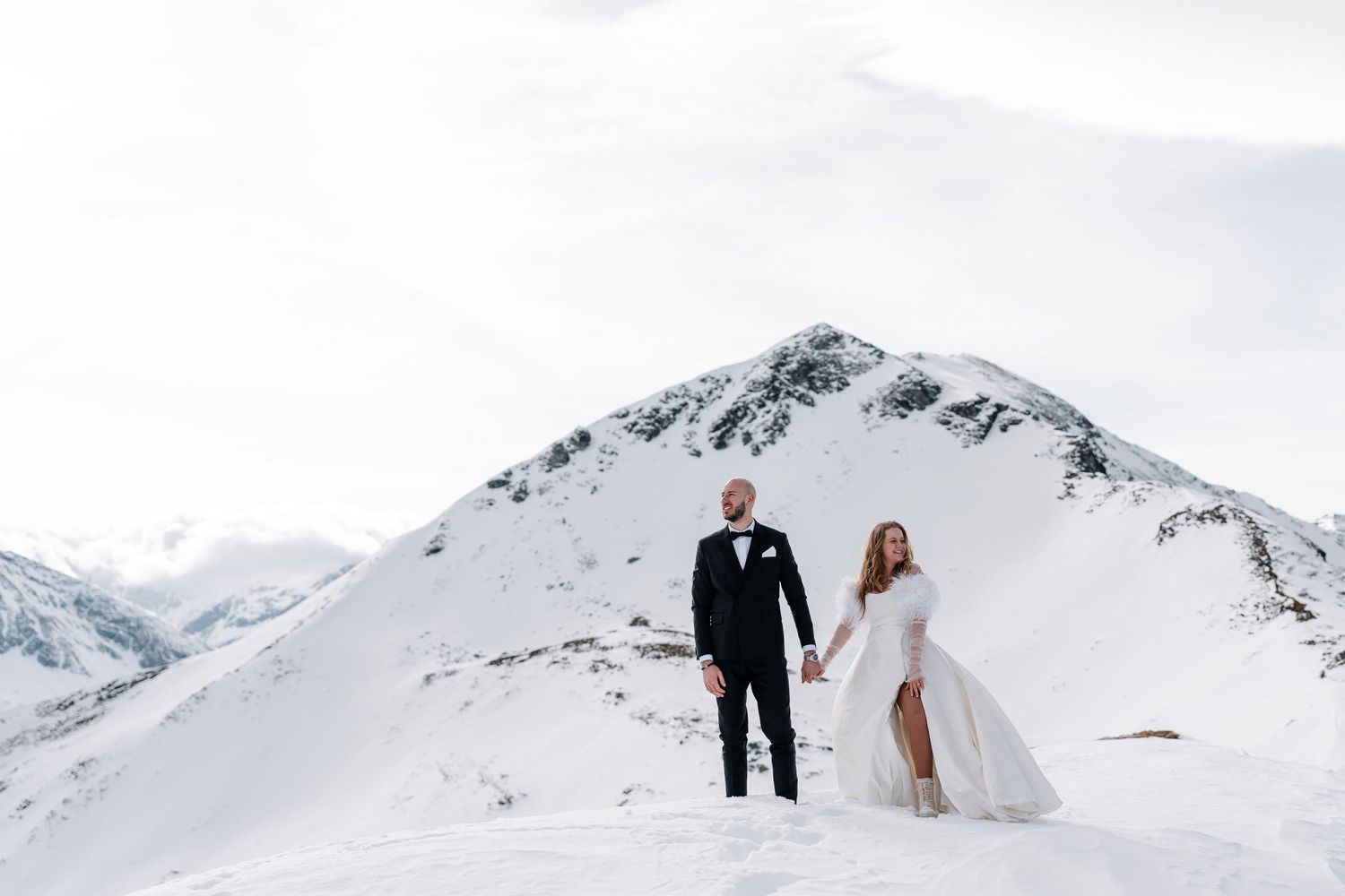 A couple in wedding attire stands atop a snowy mountain peak against a dramatic winter landscape.