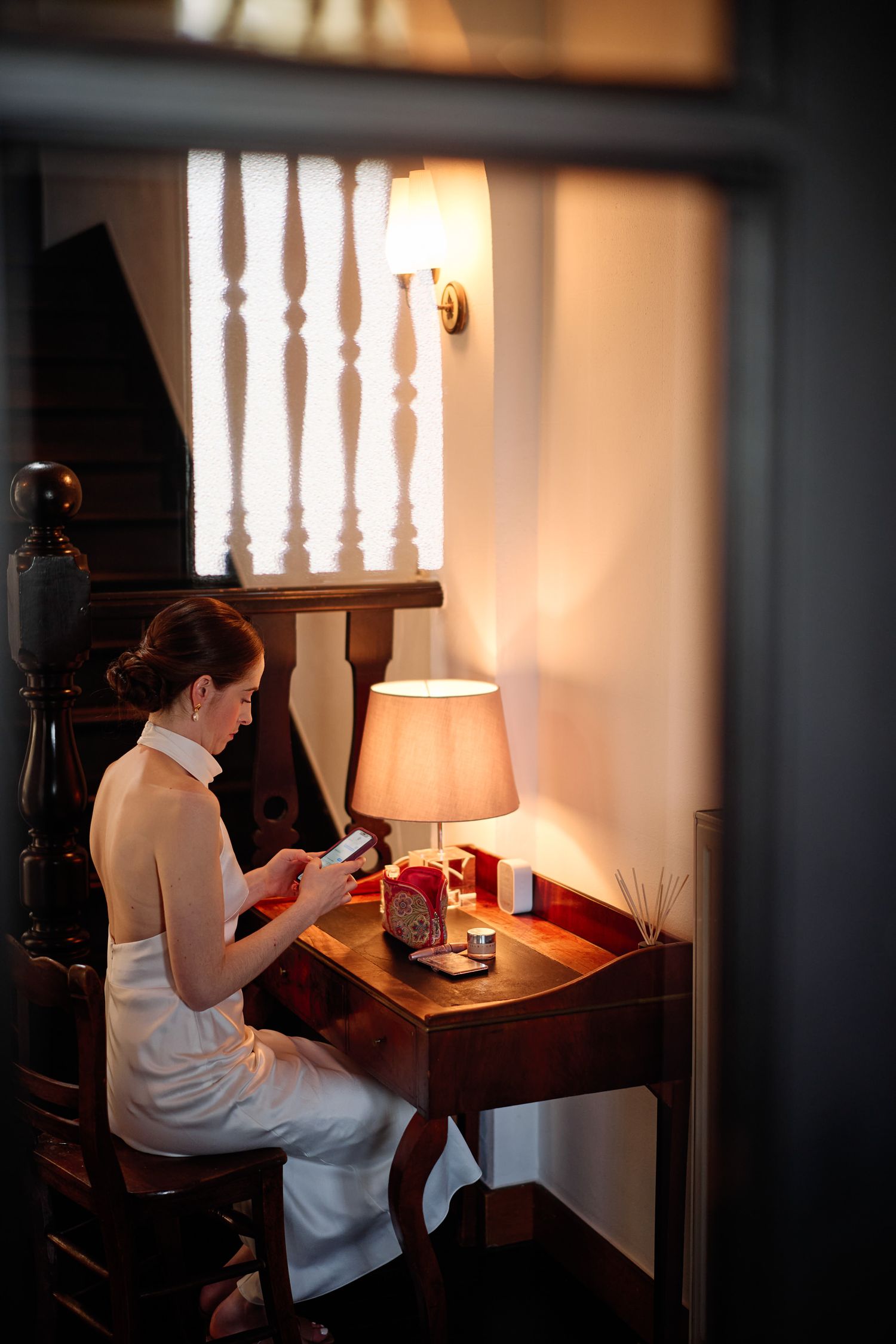 Person in white shirt sits at wooden desk with lamp reading in dark moody room with backlit window details.