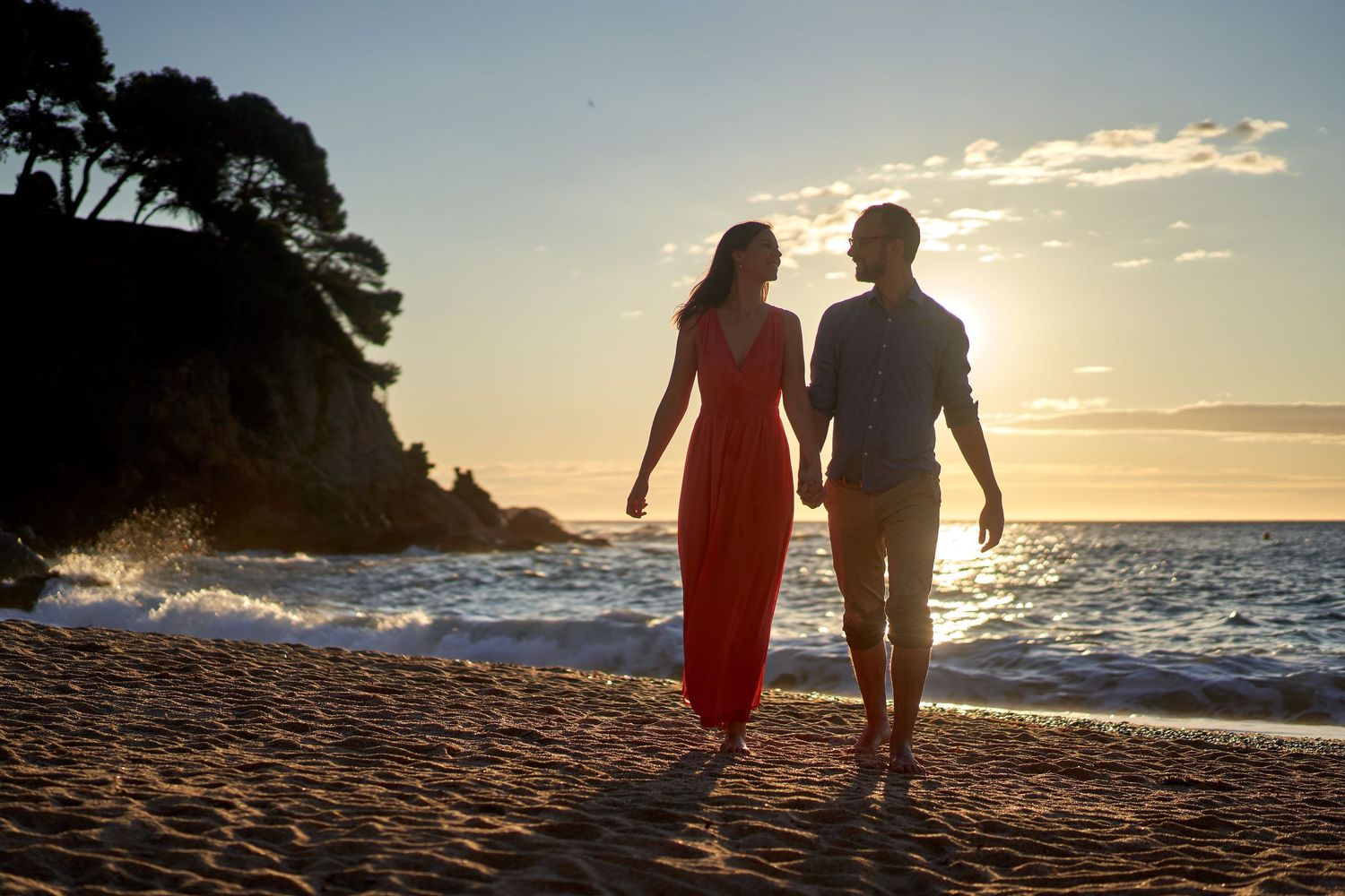 A couple walks along a scenic beach at sunset while waves crash against the shore and cliffs in the background.