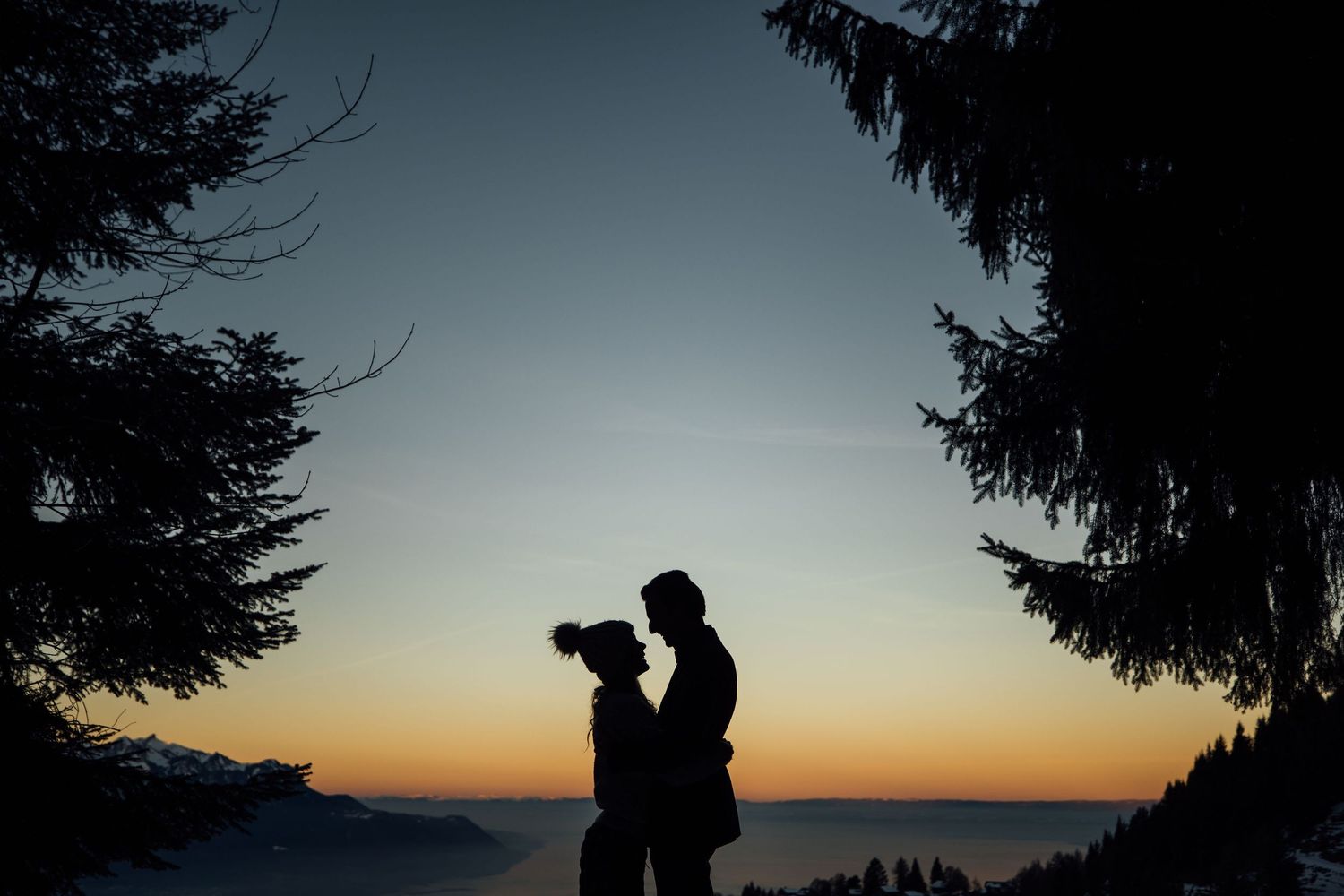 Dark silhouettes of evergreen trees frame a couple embracing against a dusky orange sunset sky.