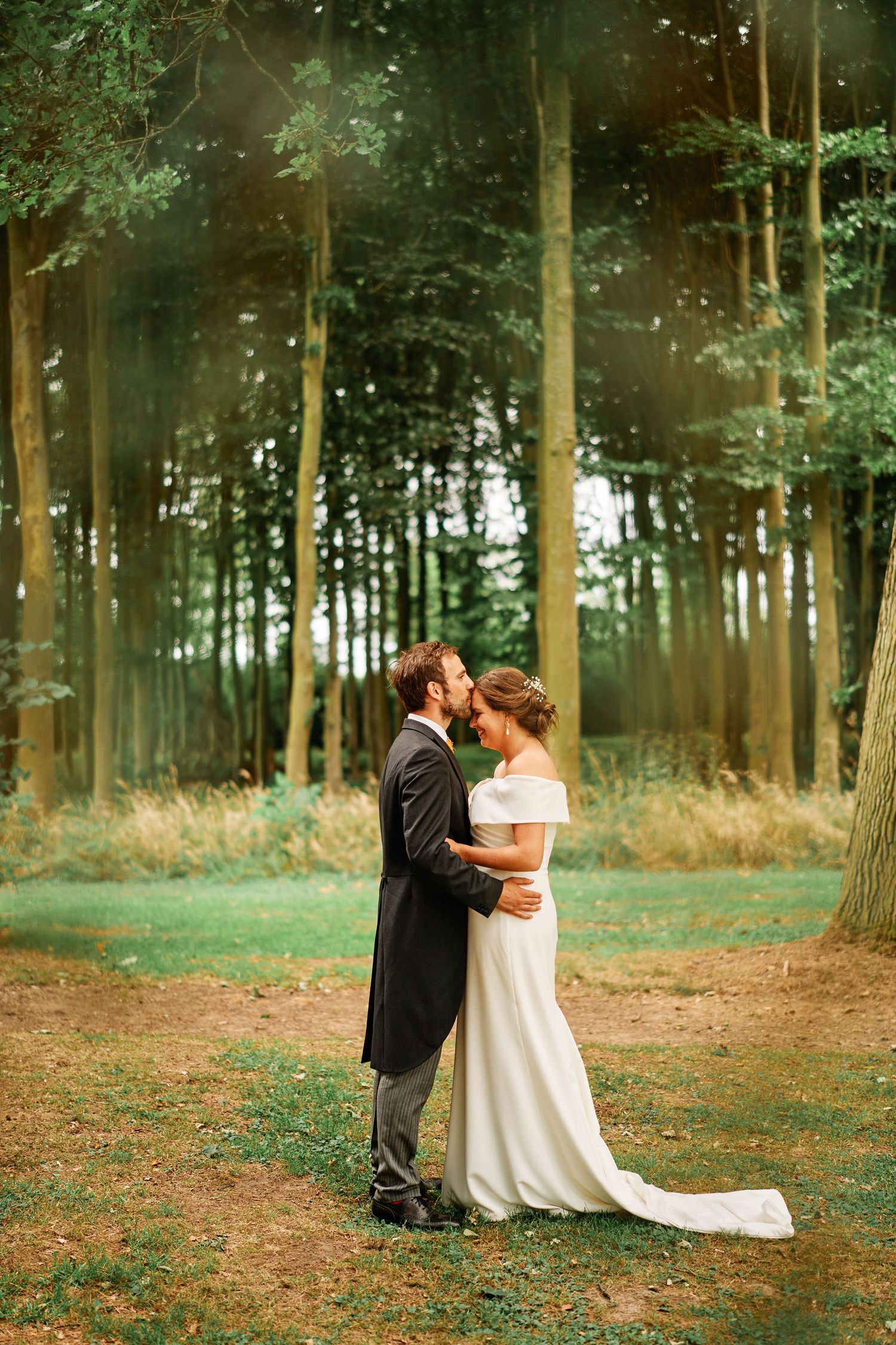 A romantic wedding couple embraces in a misty forest setting with sunlight streaming through tall trees.