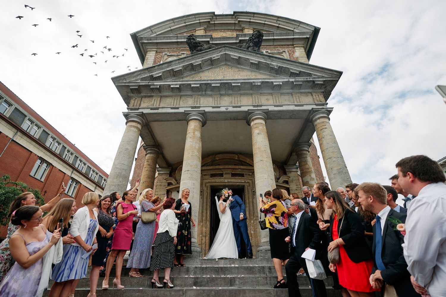 Wedding guests celebrate and dance outside a grand stone building with classical columns and architecture.