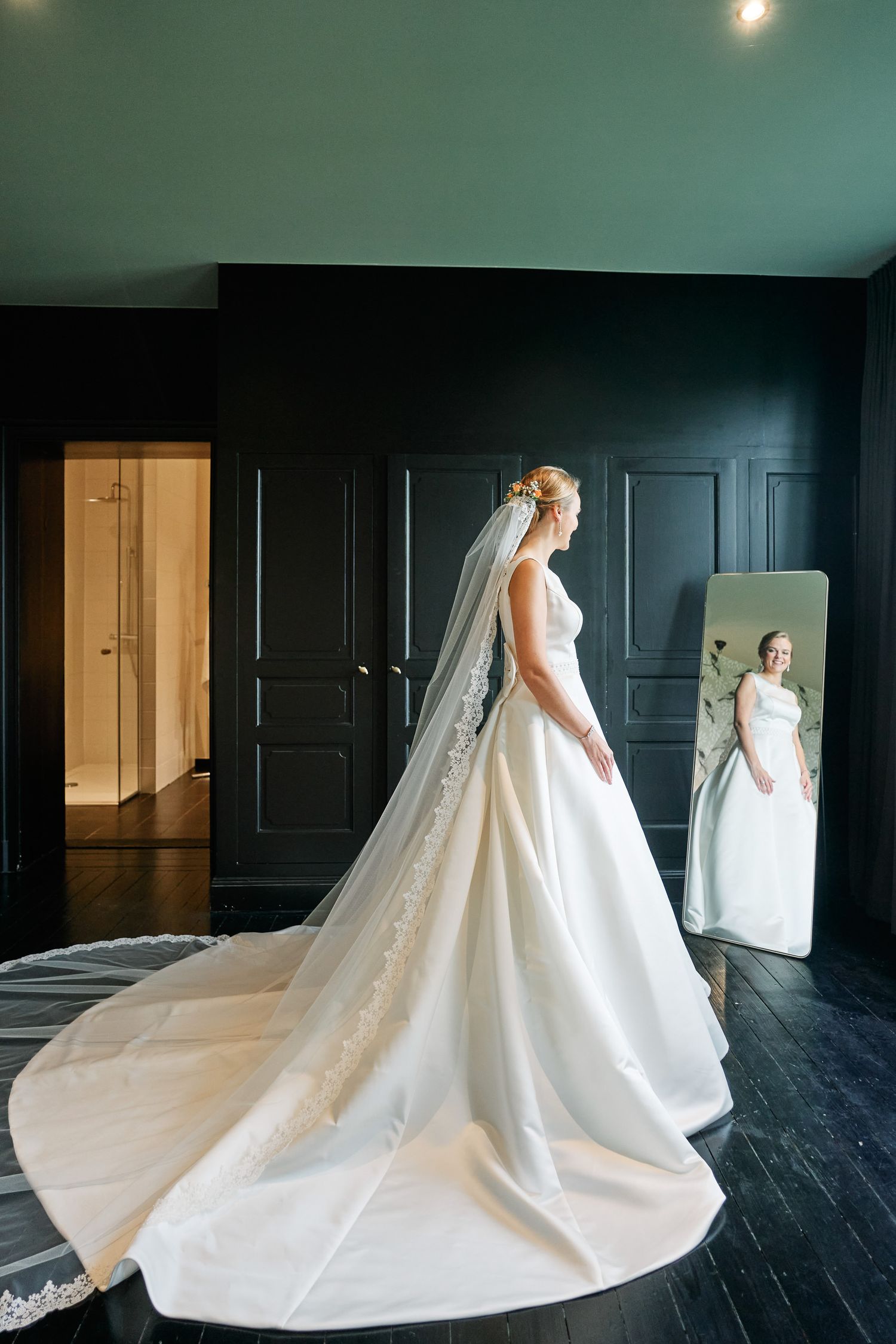 A bride in a flowing white wedding gown with a dramatic long train and veil stands before a dark wall with mirrors.