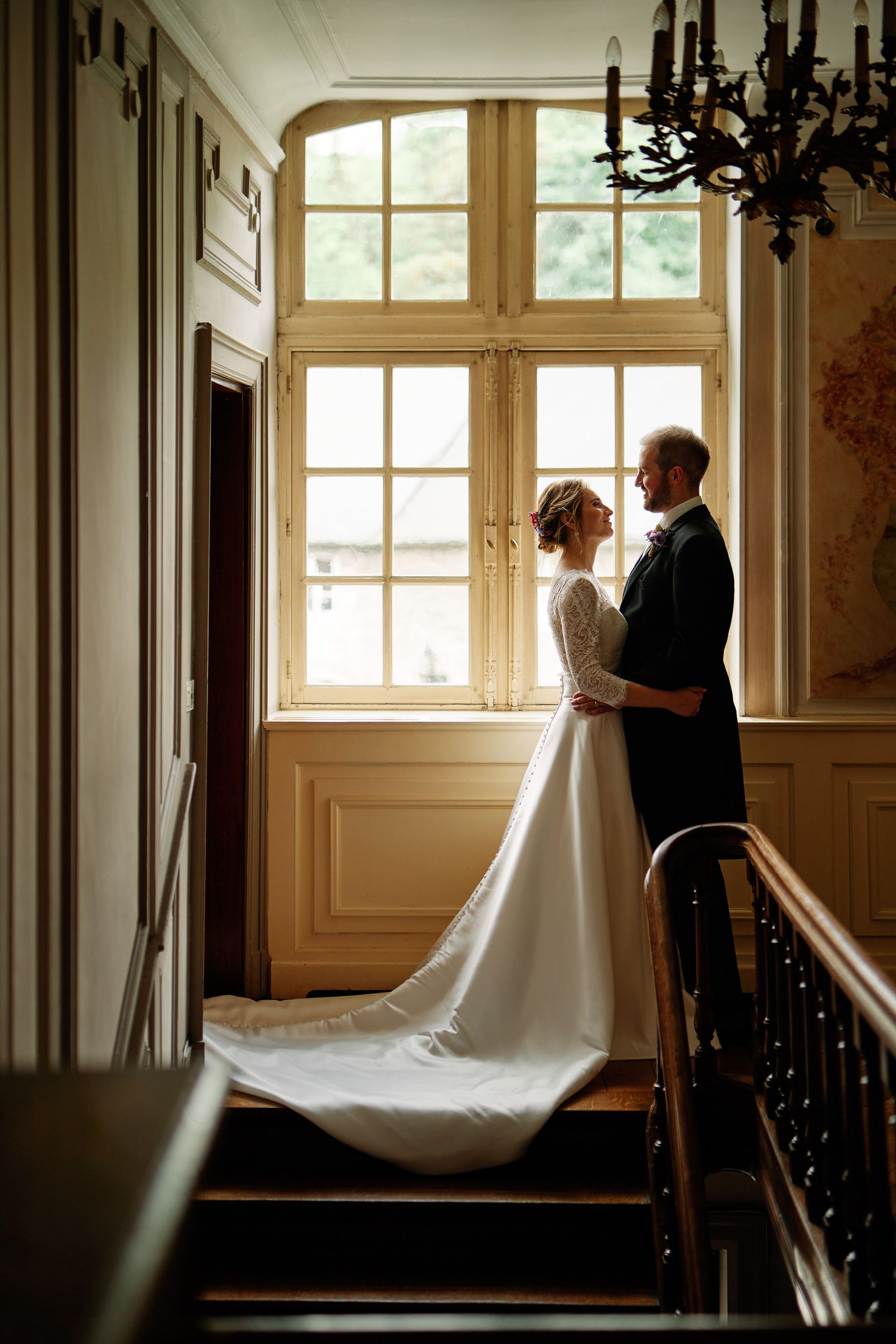 A bride and groom share an intimate moment by a window in an elegant vintage interior setting.