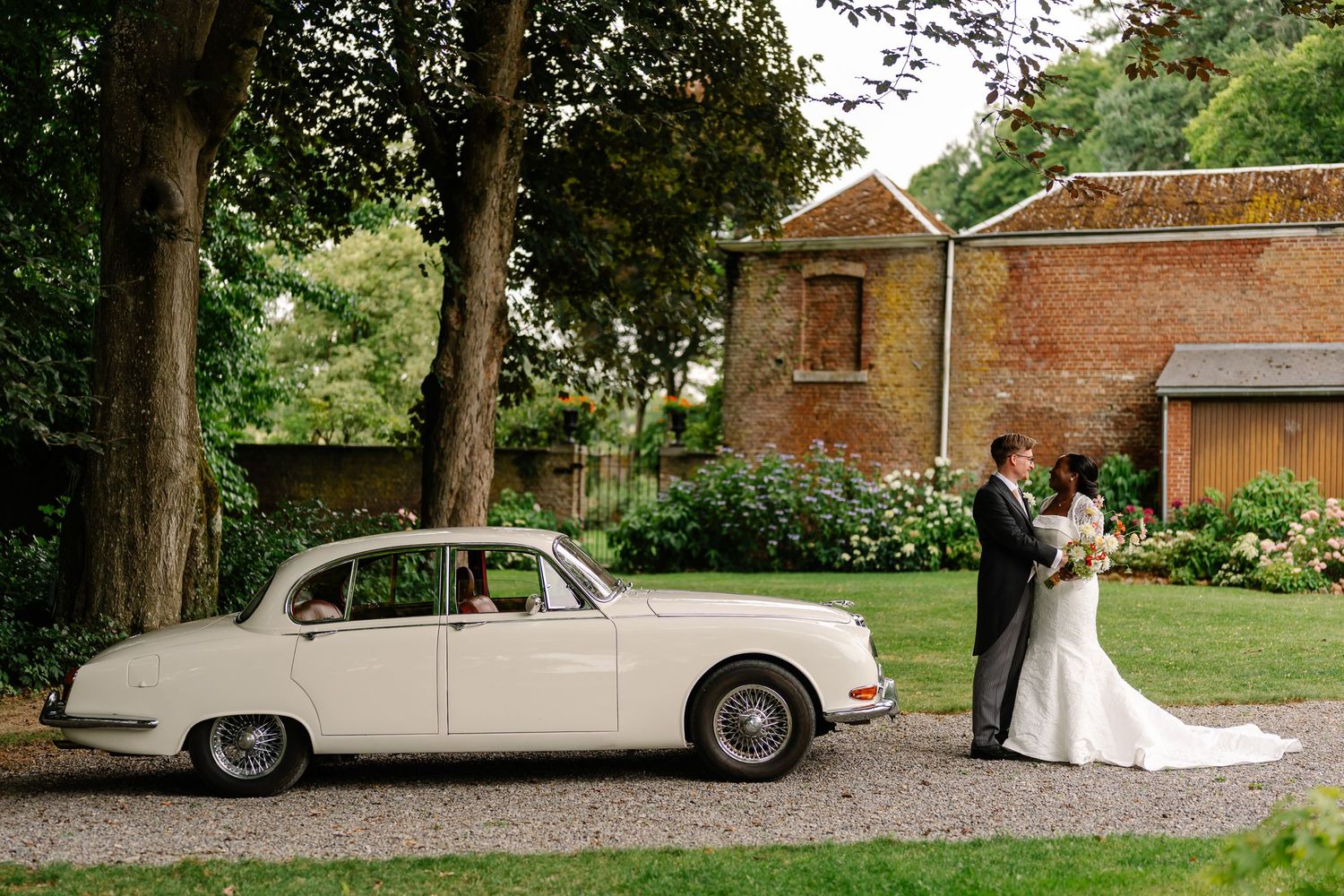Classic white vintage car parked on gravel driveway at elegant outdoor wedding venue with brick buildings.