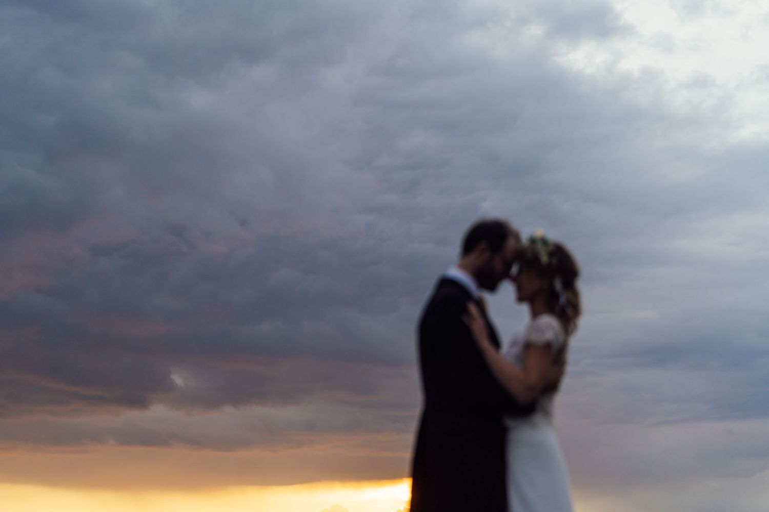 Silhouetted couple embraces against dramatic cloudy sunset sky during their wedding photoshoot.