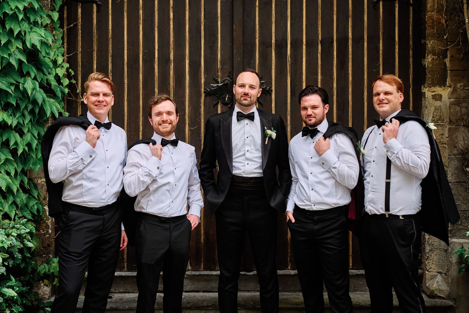 A group in formal attire stands together against a rustic wooden wall with green foliage visible.