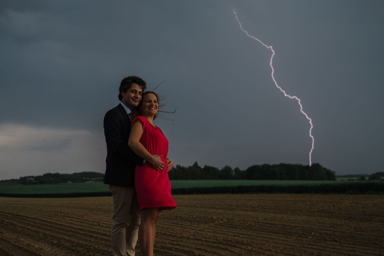 Lightning strikes dramatically illuminate the dark stormy sky over a rural field as figures embrace in silhouette.