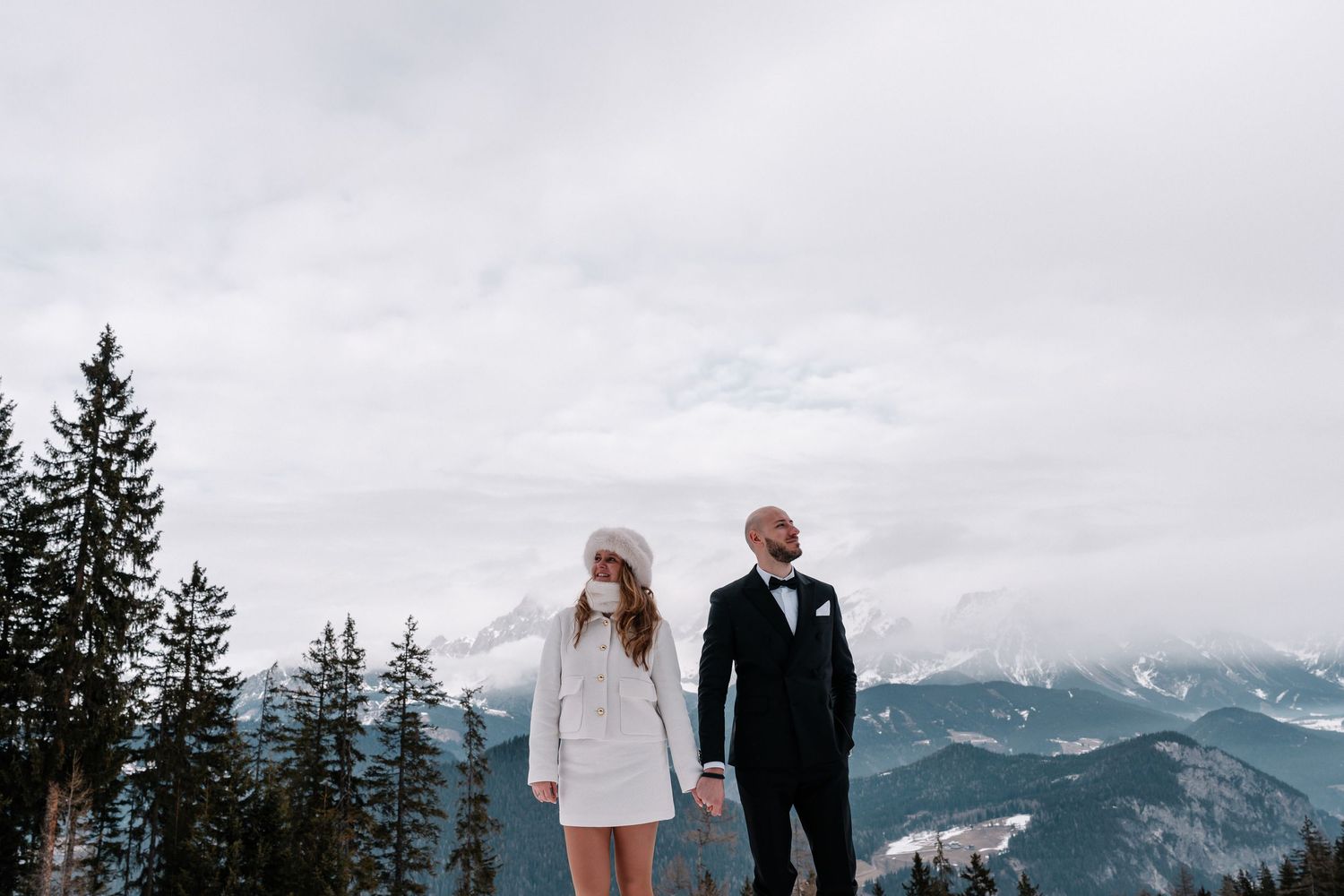 A couple stands together on a snowy mountain ridge with evergreen trees and cloudy skies in the background.