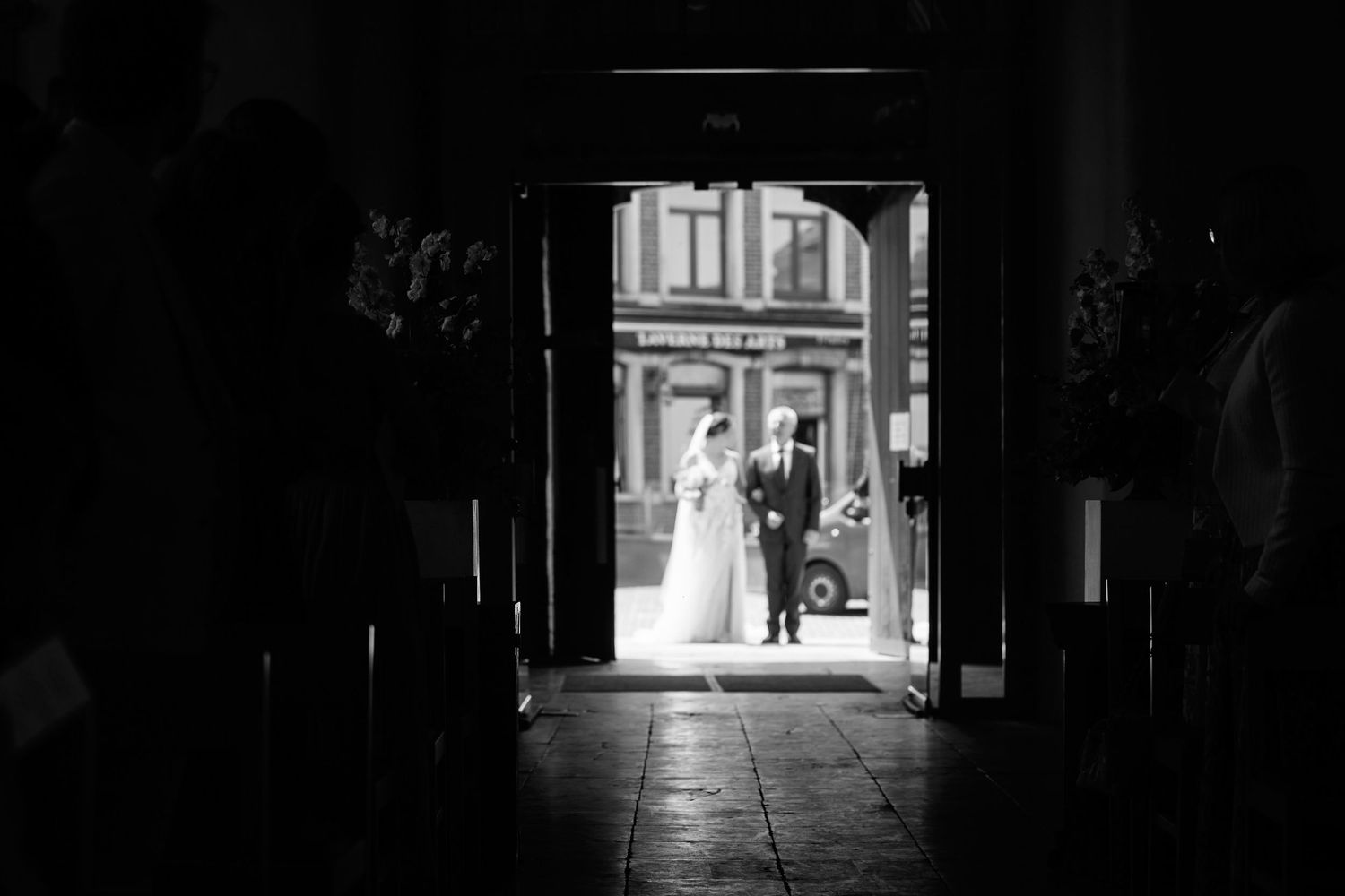 A black and white wedding photograph taken through a dark archway showing a couple in silhouette at a church entrance.