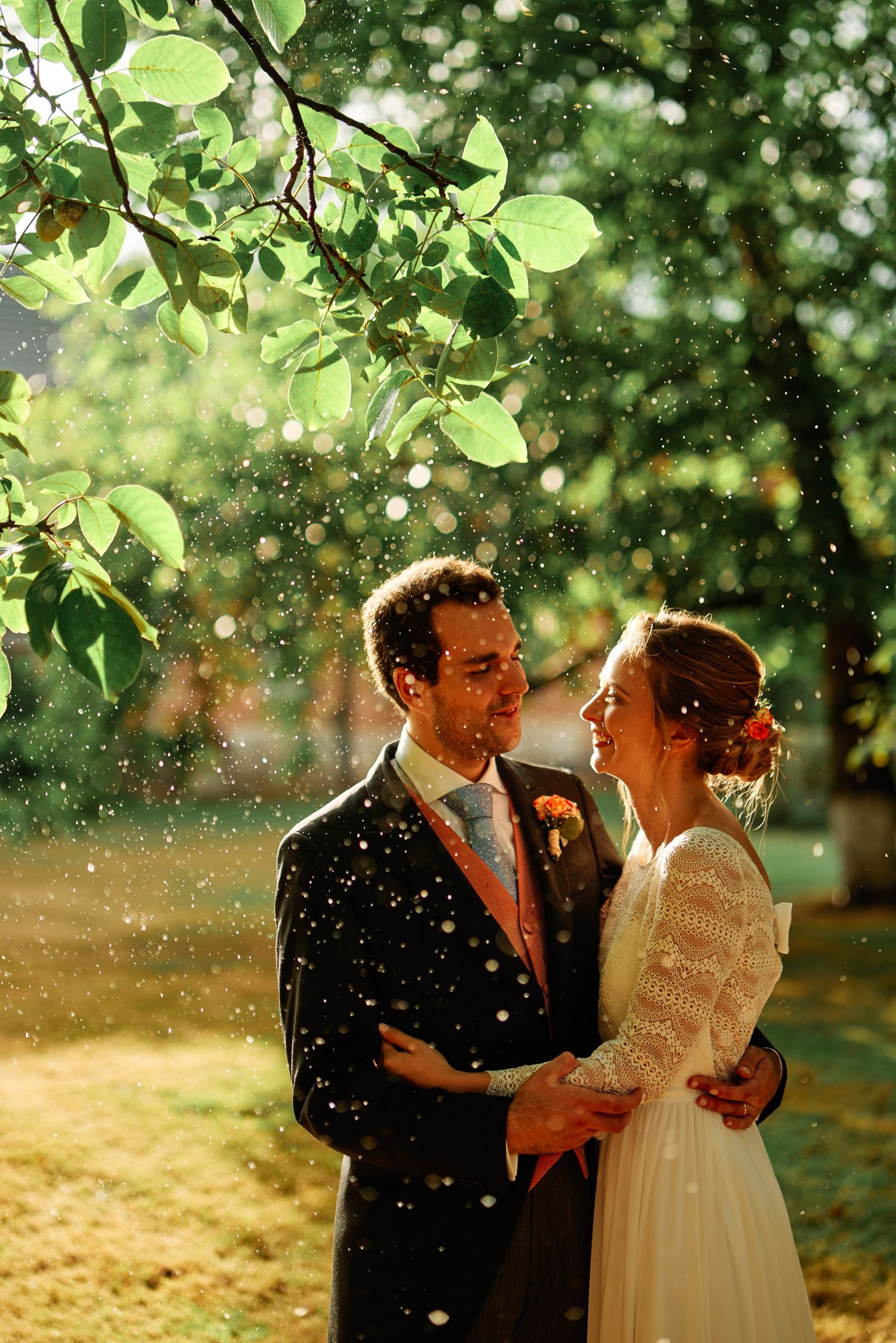 A romantic couple embraces in a dreamy garden setting with sunlit bokeh and green foliage in the background.