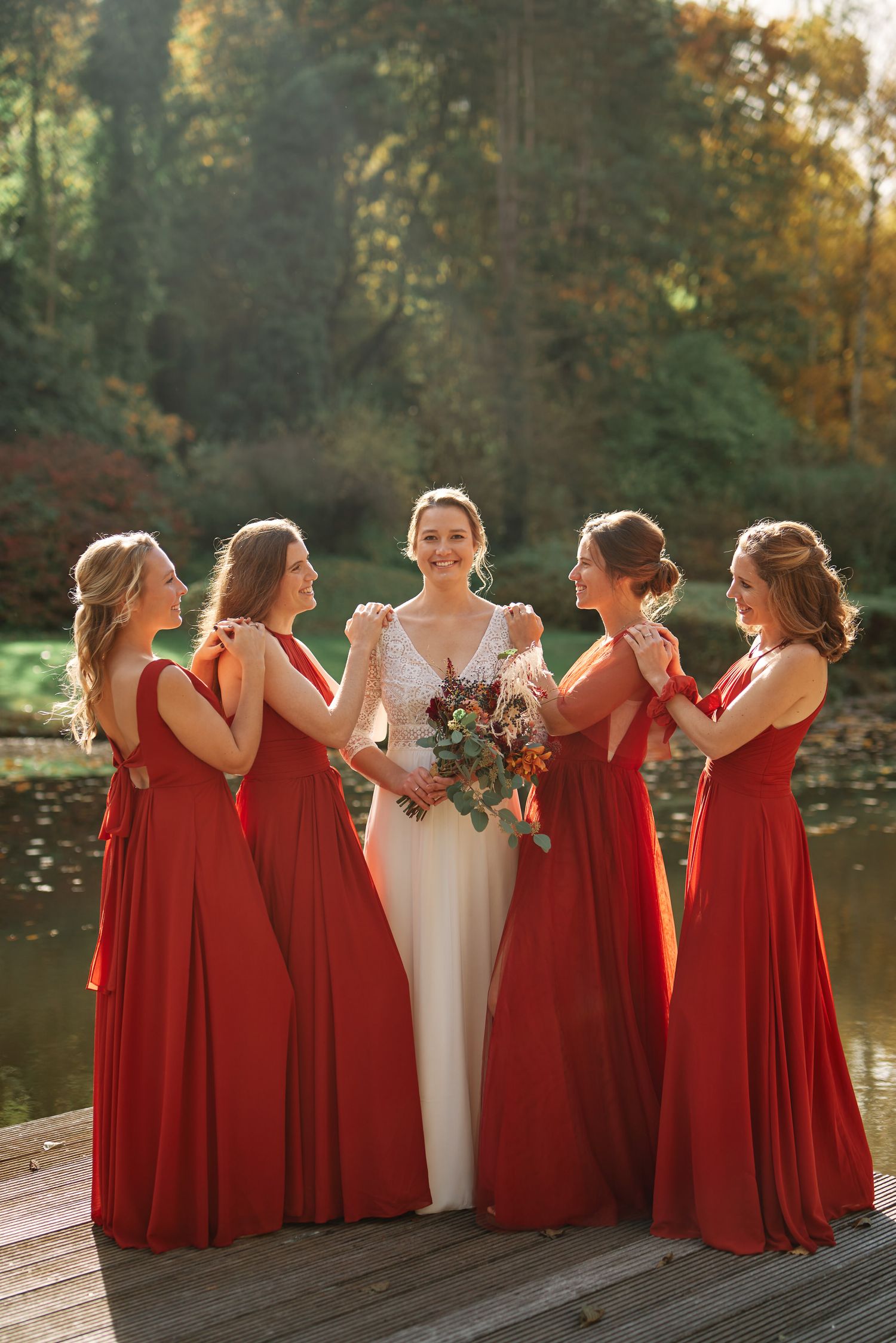 A bridal party in deep red dresses stands together on a dock by a lake during golden hour.