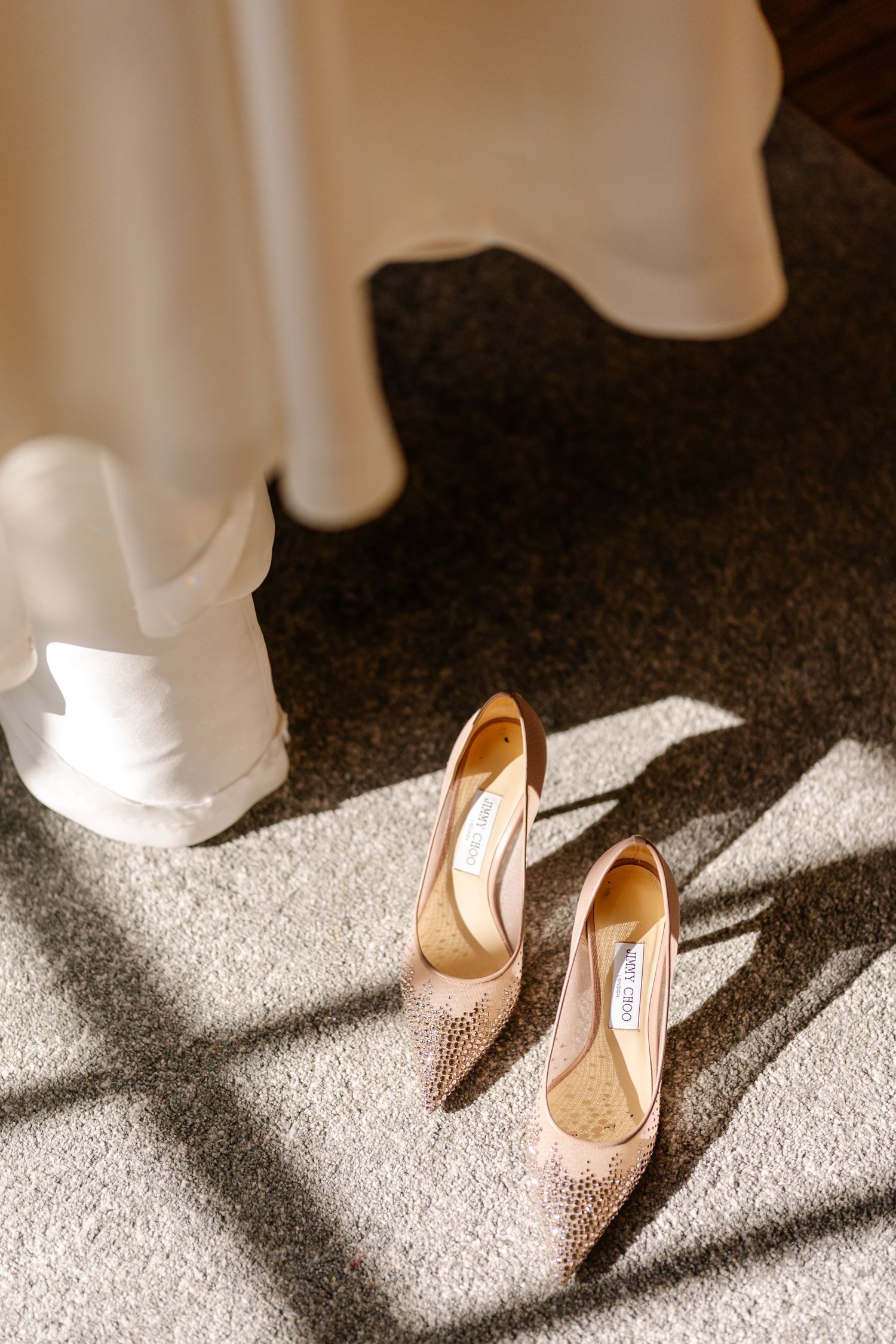 Elegant nude heels cast dramatic shadows on a light carpet while a white dress hem flows above them.