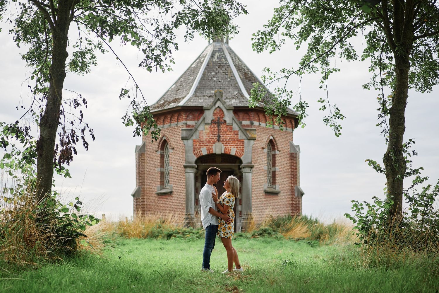 A couple shares a romantic moment near a rustic stone chapel surrounded by lush trees and greenery.