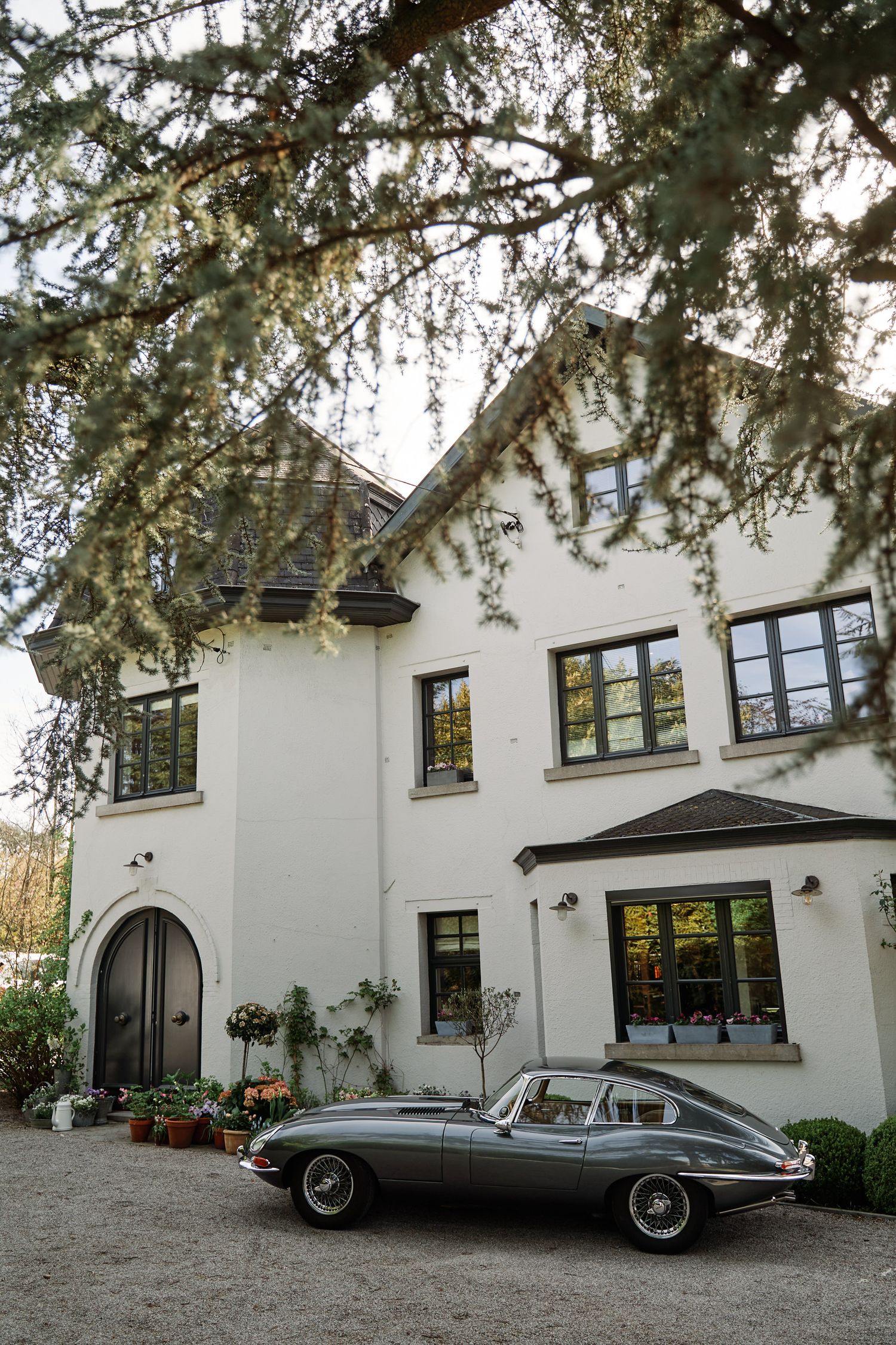 Classic white mansion with black-trimmed windows and vintage car parked outside in driveway under tree branches.