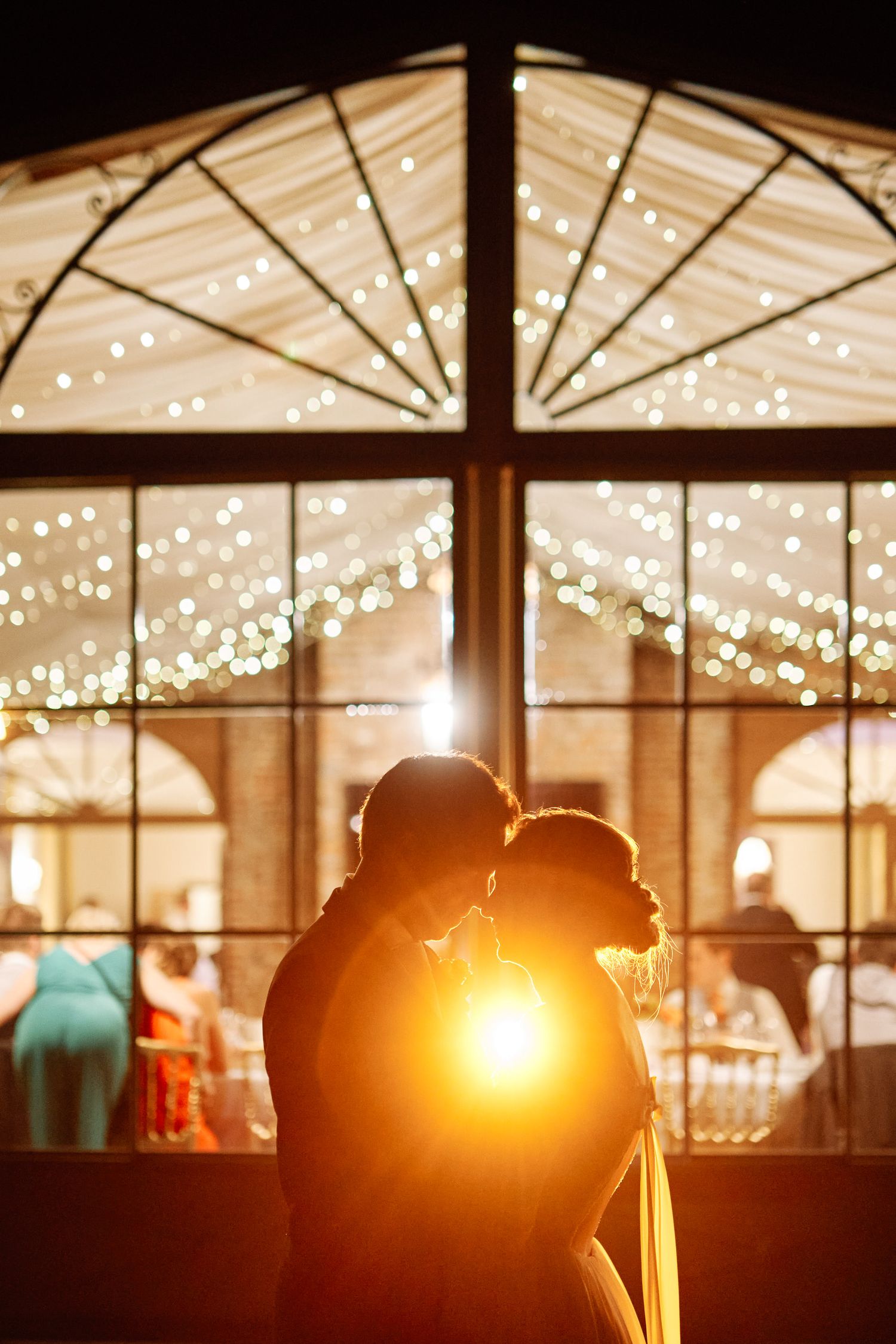 Silhouettes of people dancing under warm string lights in a wedding reception venue at sunset.