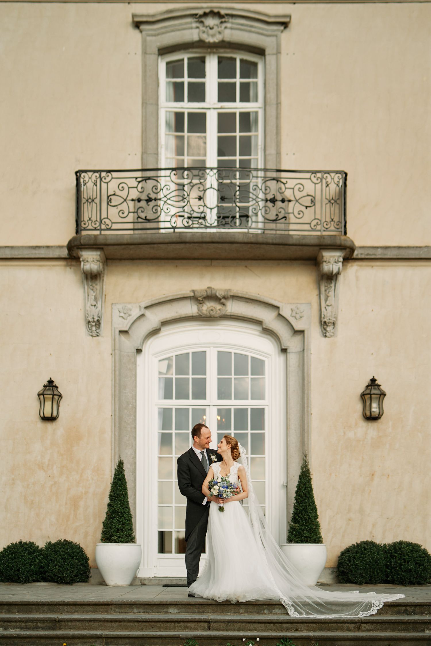 Elegant wedding couple poses in front of a classic mansion with arched windows and ornate balconies.