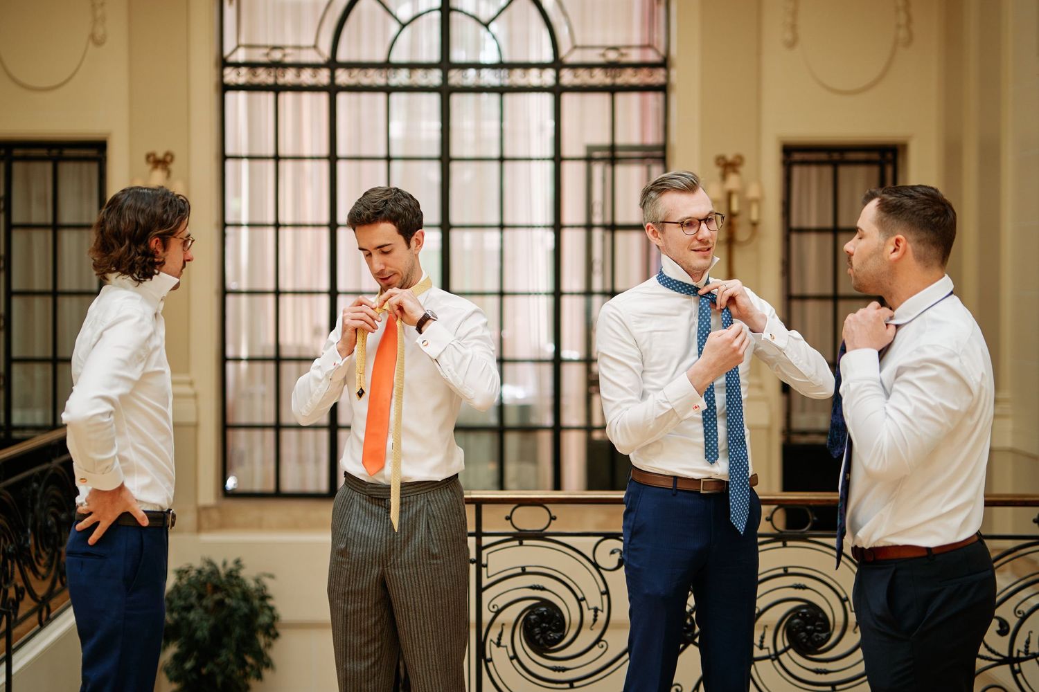 Groomsmen in white shirts and navy suits prepare for a wedding in an elegant hallway with arched windows.