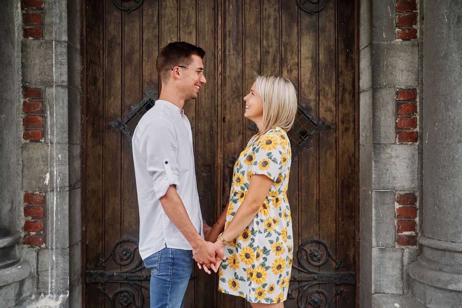 Couple stands in front of rustic wooden barn door holding hands during engagement photo session.