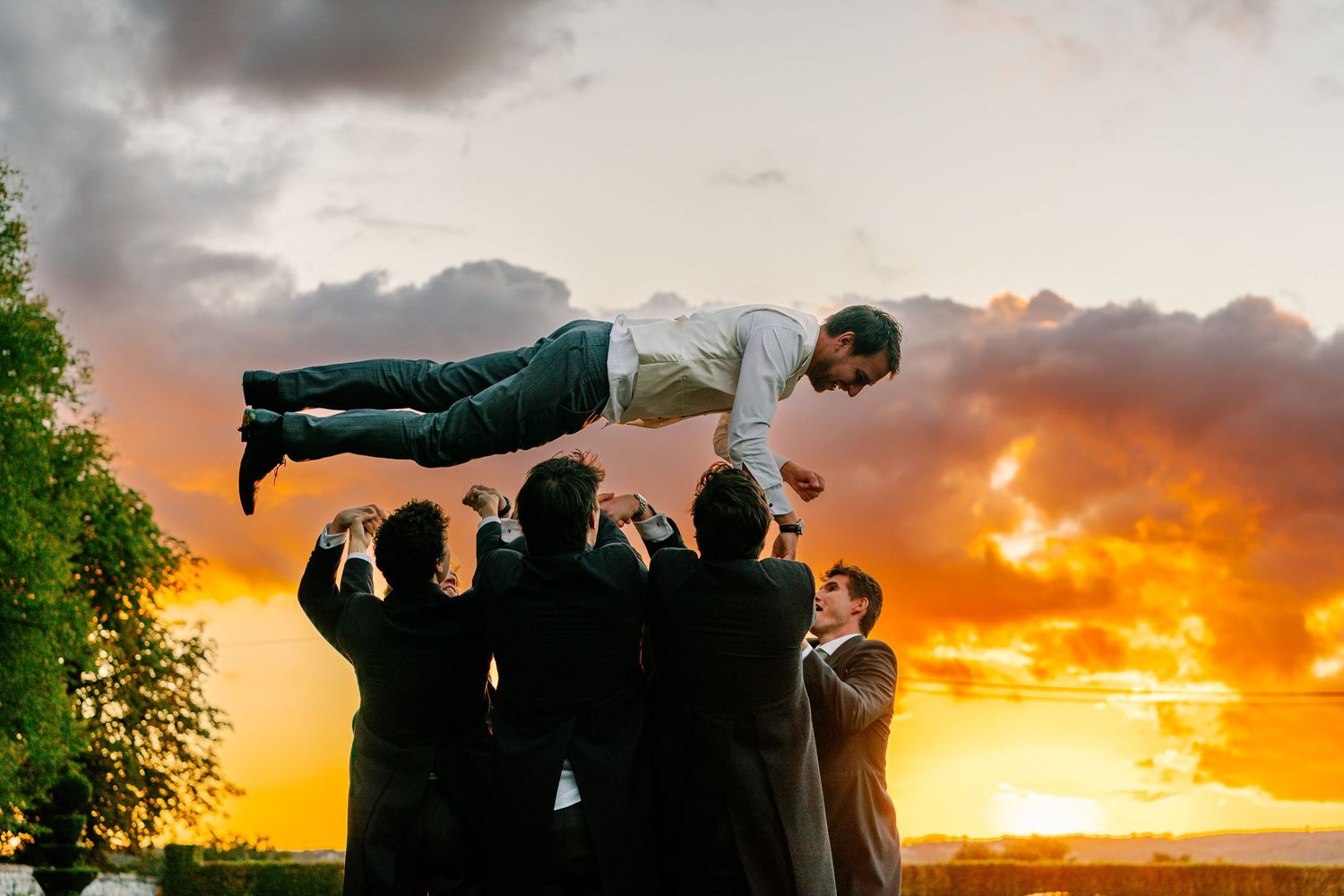 Groomsmen lift and hold a person horizontally against a dramatic sunset sky with orange clouds.