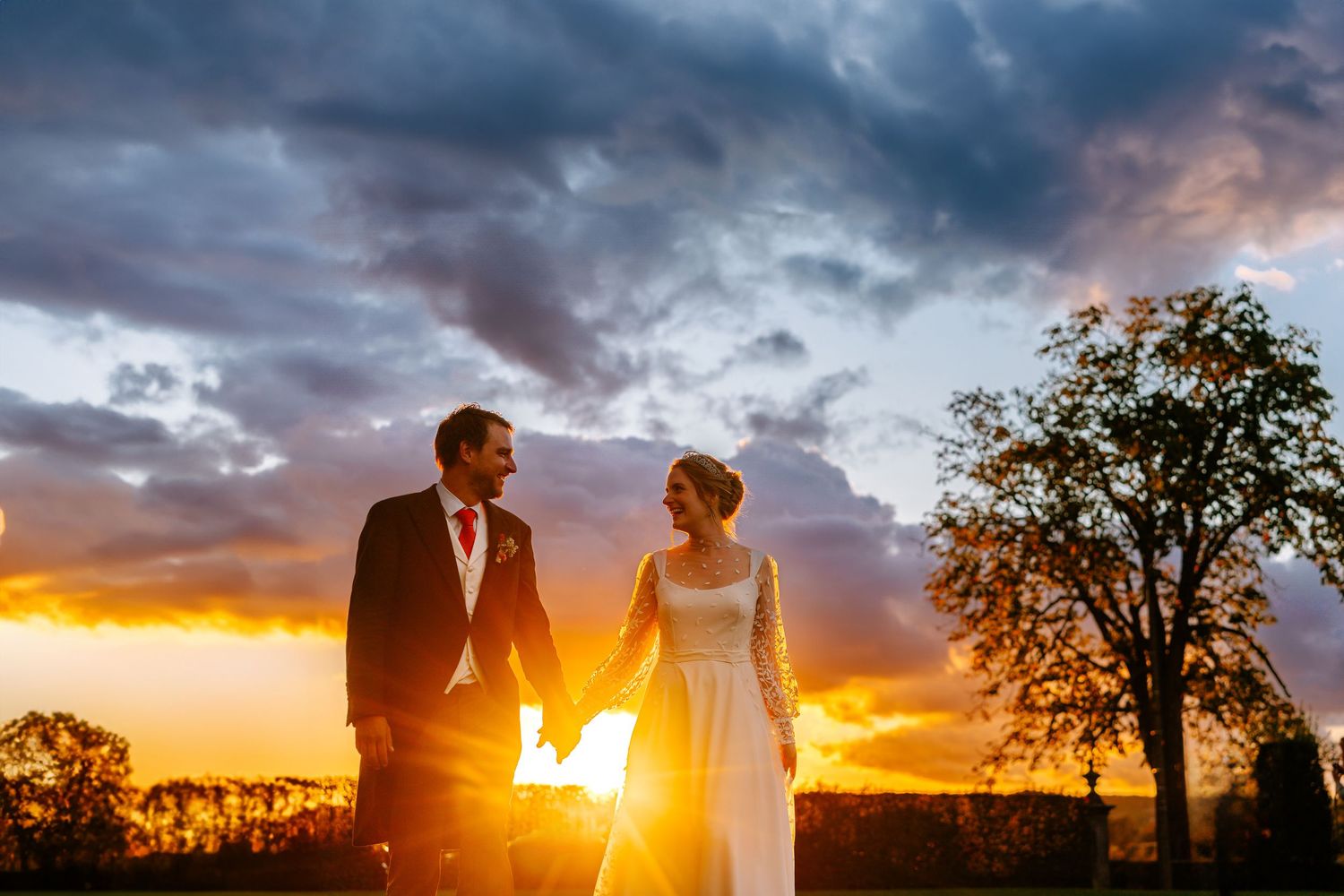 A couple stands silhouetted against a dramatic sunset sky with trees in the background during their wedding photoshoot.