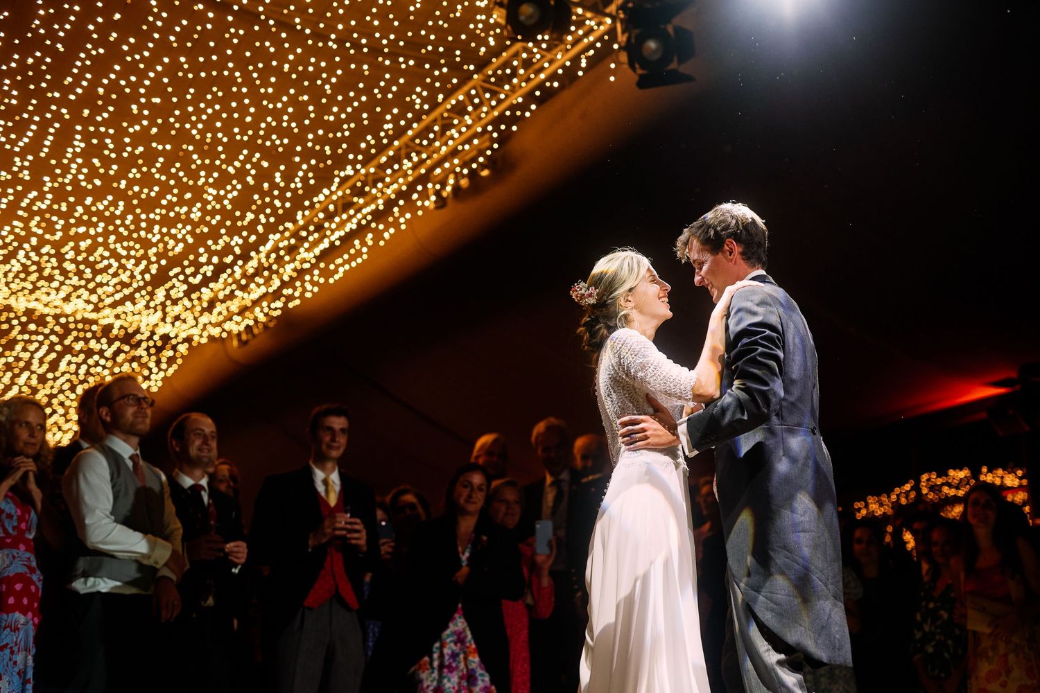 A couple shares their first dance at a wedding reception under a canopy of warm glowing string lights.