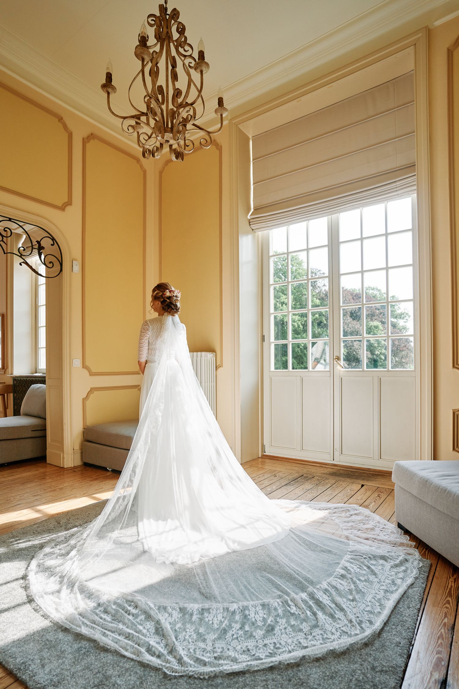 A flowing white wedding veil with intricate lace details spreads across a hardwood floor in an elegant room with chandeliers.