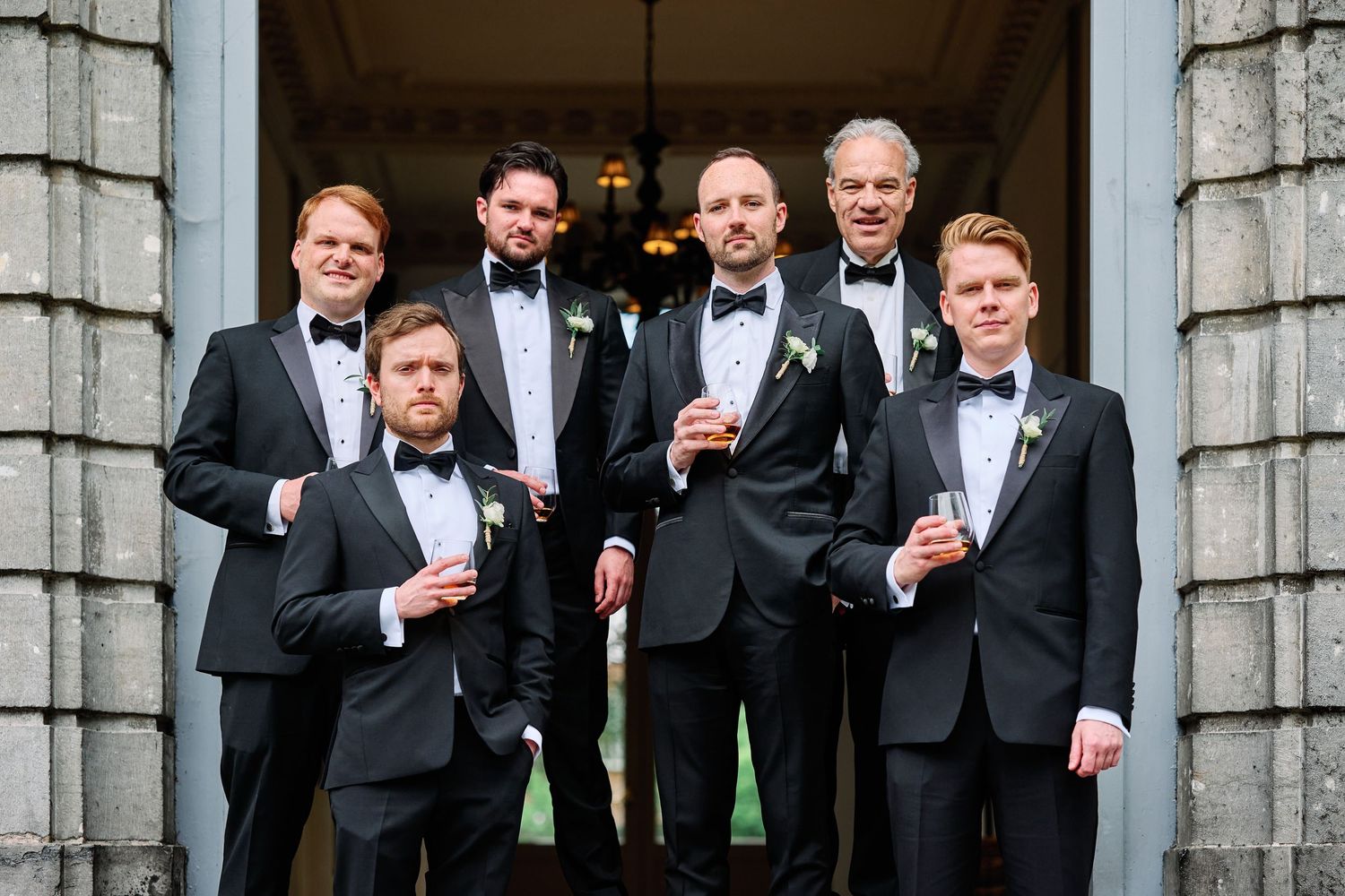 Groomsmen in black tuxedos pose together while holding drinks in front of a stone building's columns.