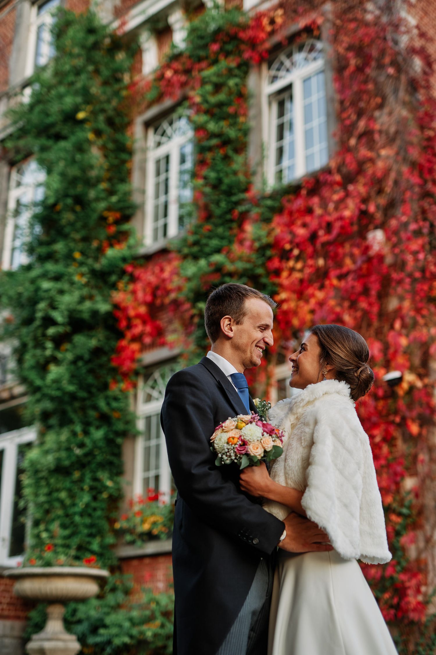A couple shares a romantic moment in front of a brick building with vibrant red autumn ivy climbing the walls.