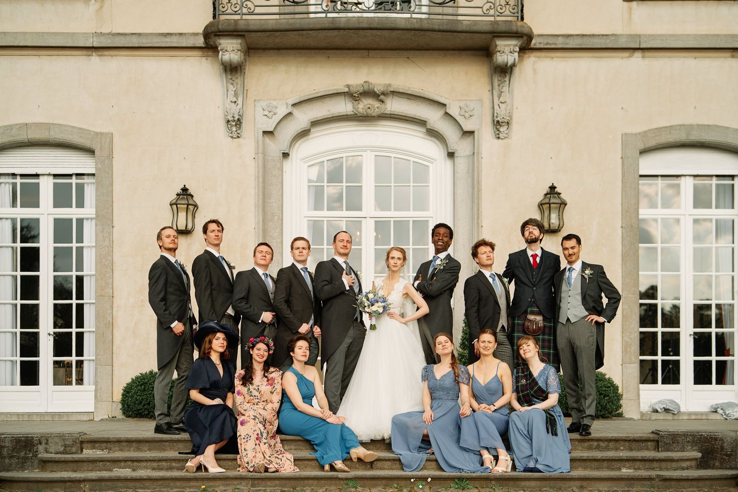 Wedding party poses together in formal attire on the steps of an elegant stone building with arched windows.