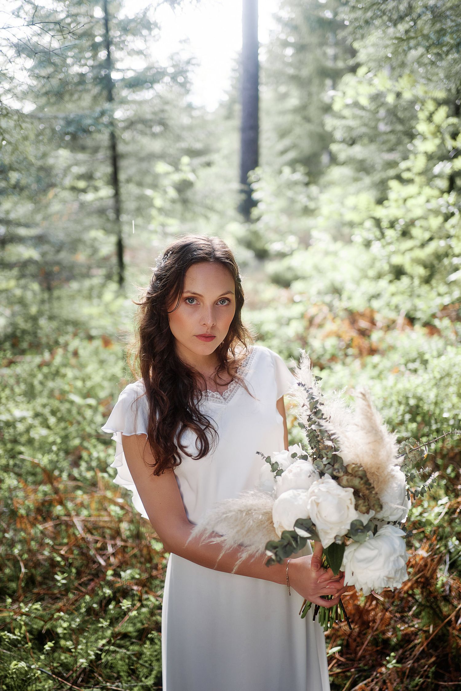 A person in a white dress poses with a bouquet in a lush forest setting with sunlight filtering through the trees.