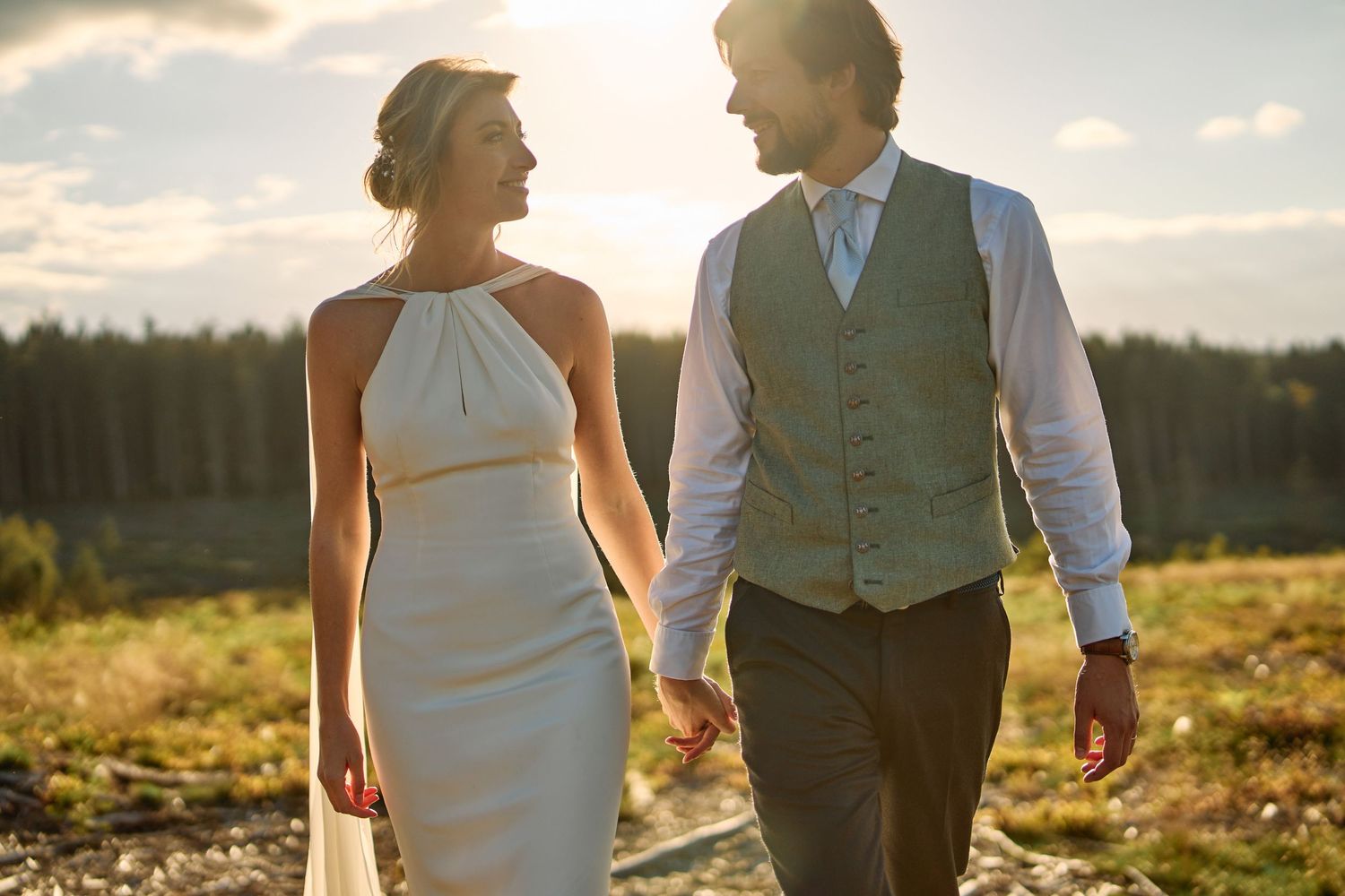 Couple in wedding attire walks through golden field at sunset holding hands during outdoor countryside photoshoot.