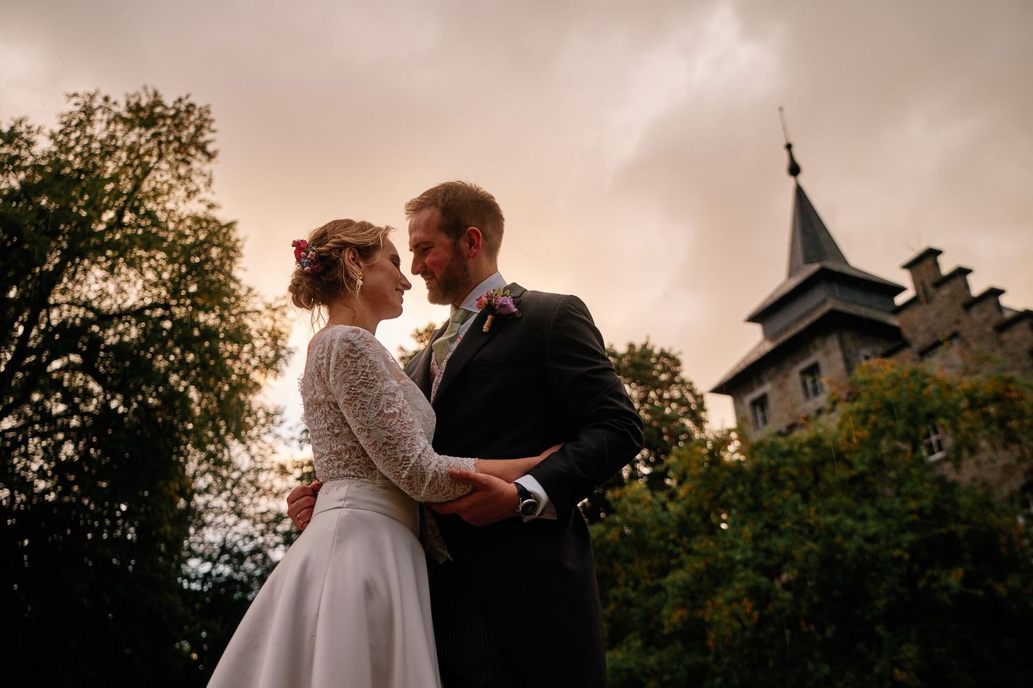 A romantic wedding couple shares an intimate moment at sunset with a castle-like building in the background.