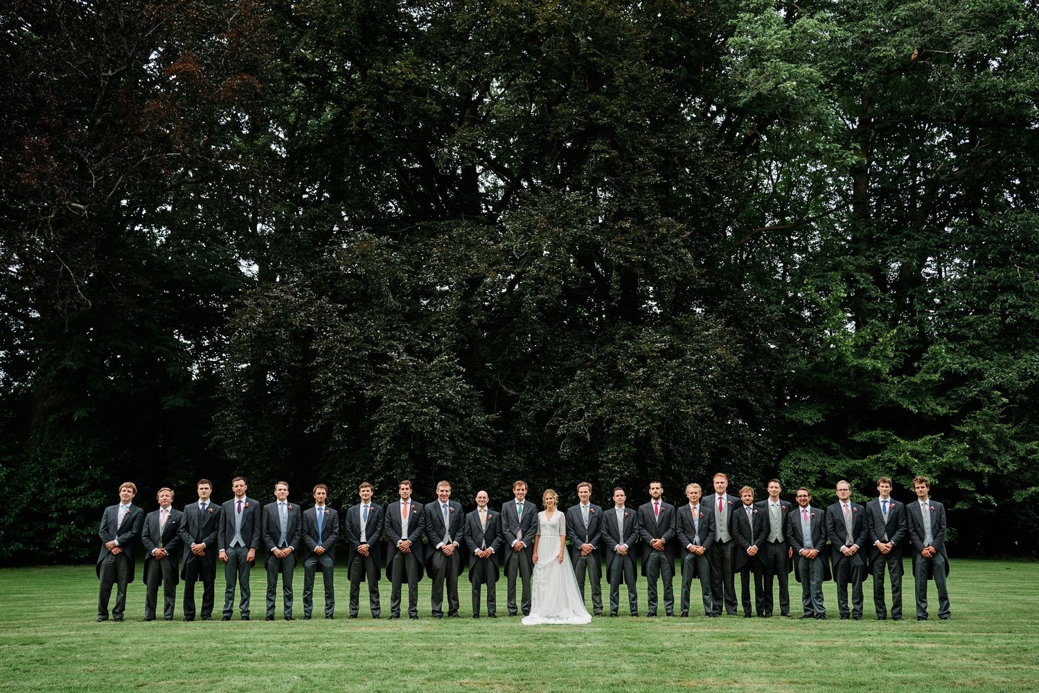 A wedding party in formal attire stands in a line formation on a lush green lawn surrounded by trees.