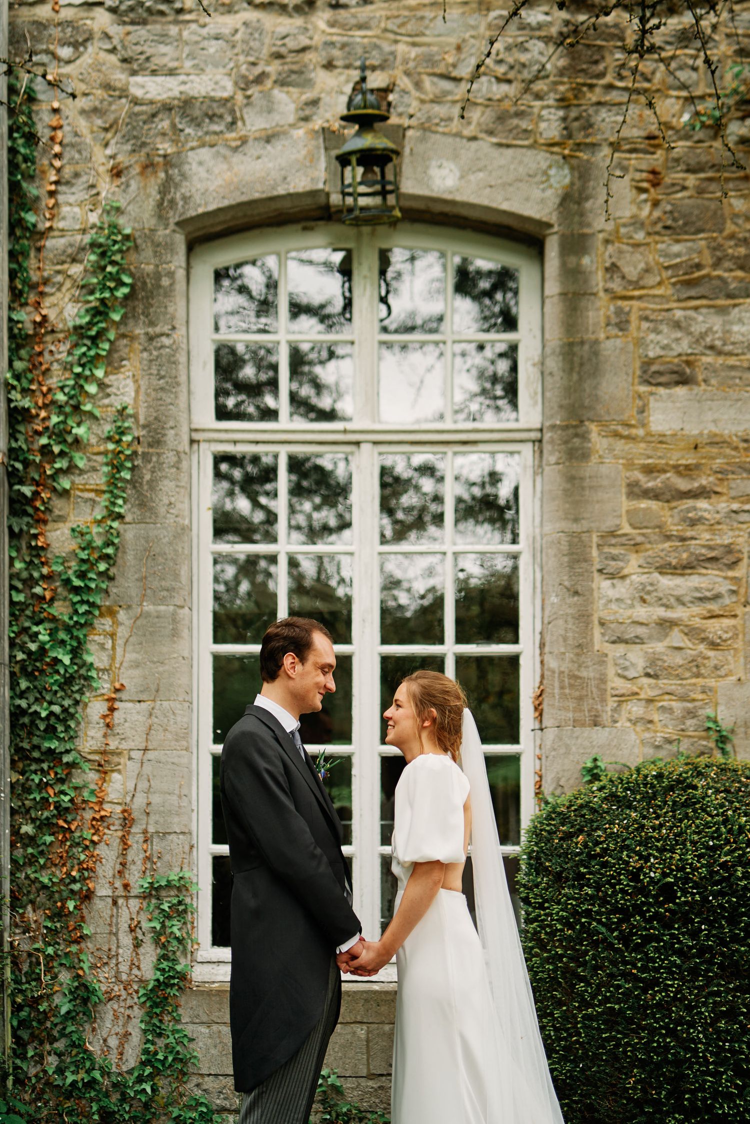 A wedding couple stands by a historic stone building with ivy and arched windows during their intimate outdoor ceremony.