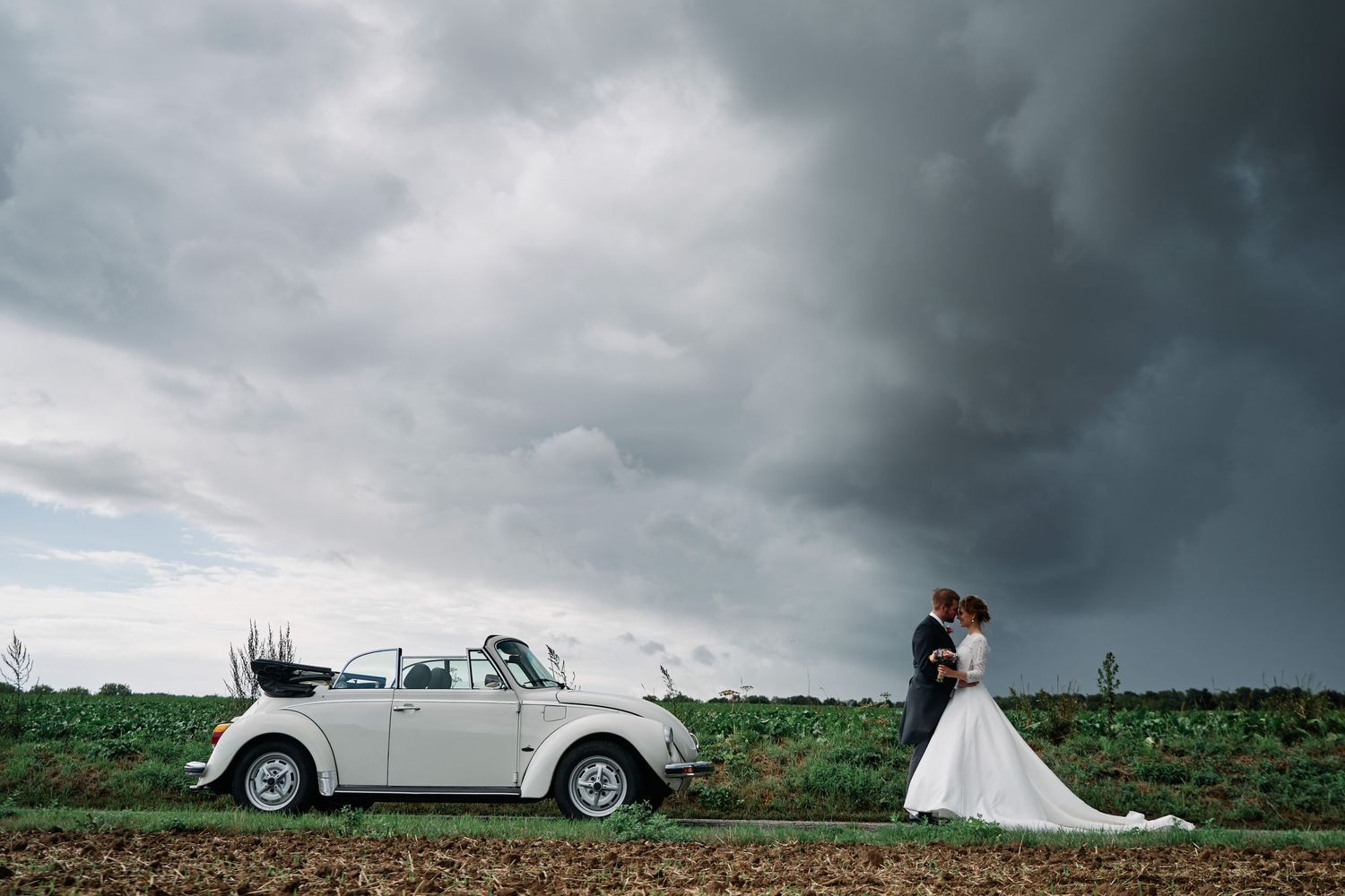 Storm clouds create dramatic backdrop for outdoor wedding photos with vintage car and flowing white dress.