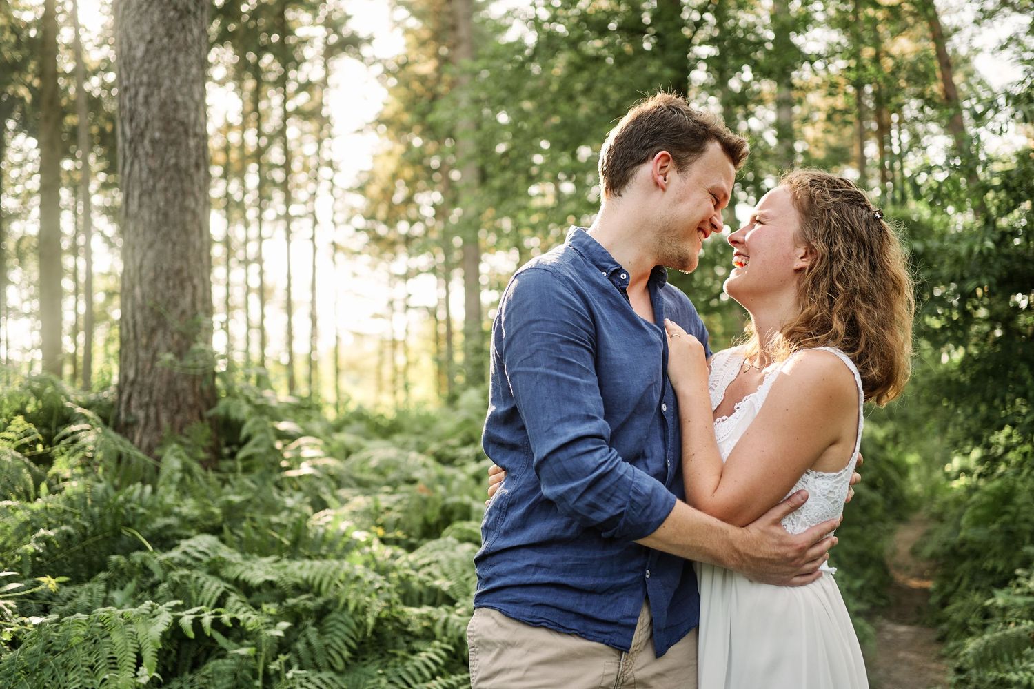A couple shares a romantic moment in a sunlit forest with tall evergreen trees in the background.